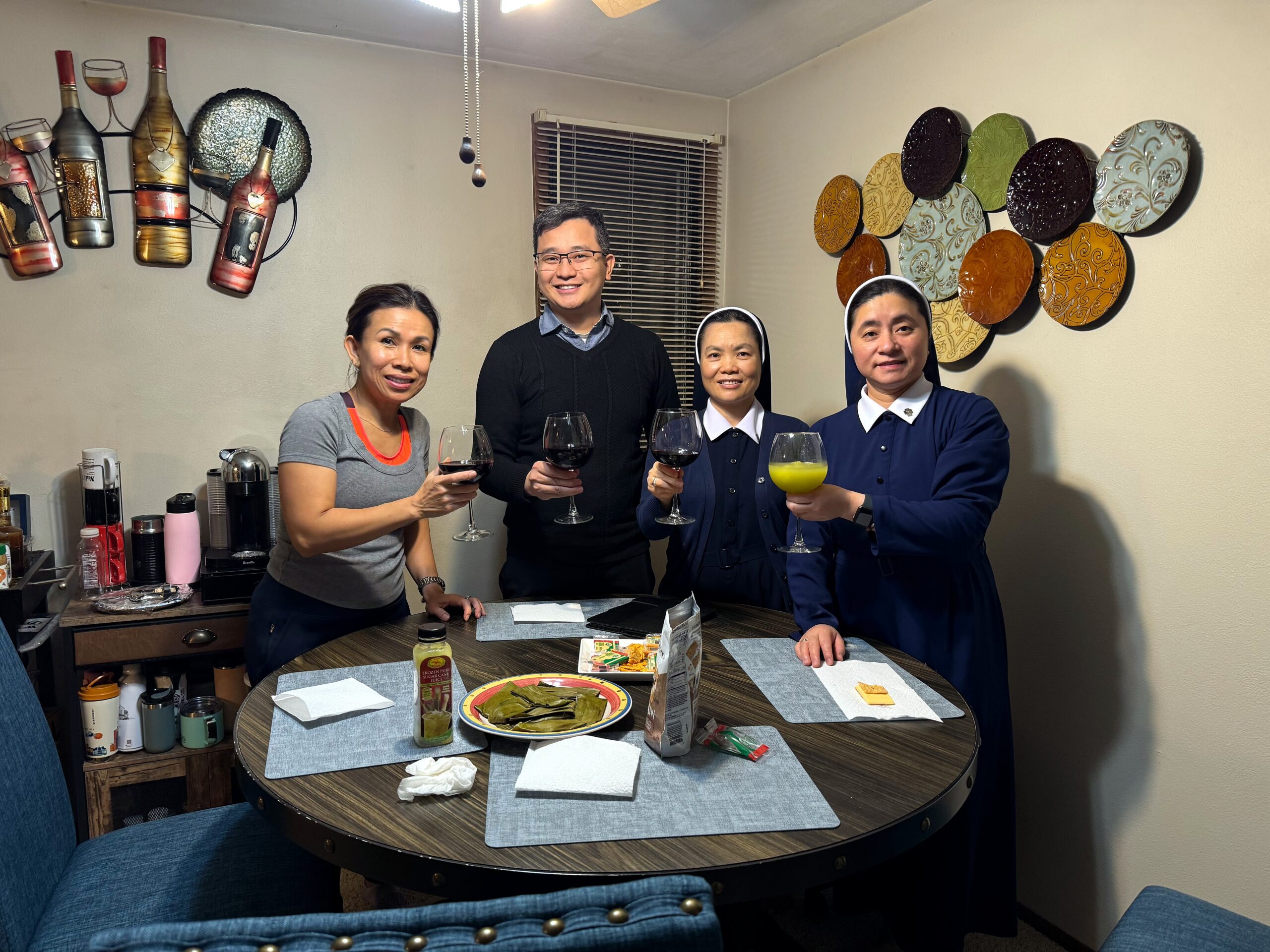 Four people stand around a round dining table, holding wine glasses and smiling in a cozy room with bottle wall decor.