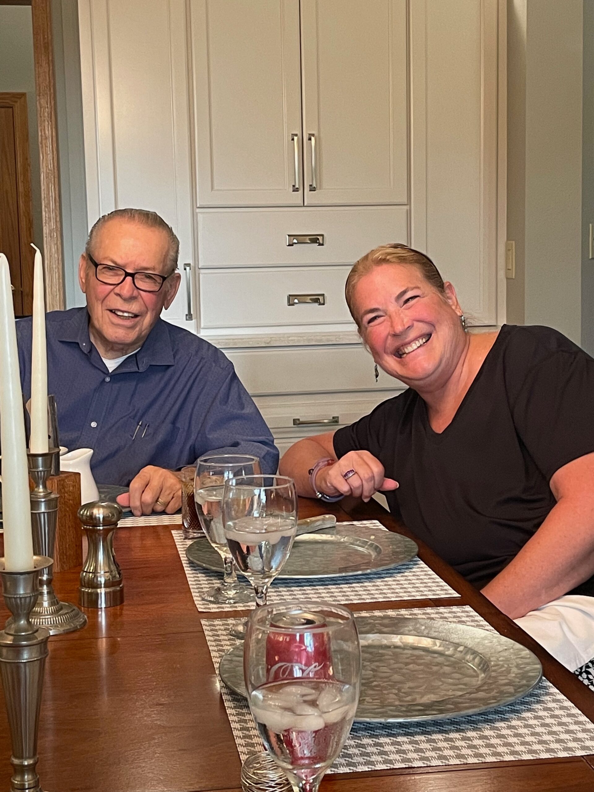 Two smiling adults sit at a dining table with candles, dishes, and wine glasses, in a kitchen with white cabinets behind them.