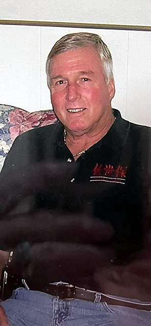 Older man with gray hair sits on a floral sofa, wearing a black polo shirt with a red logo on the chest.