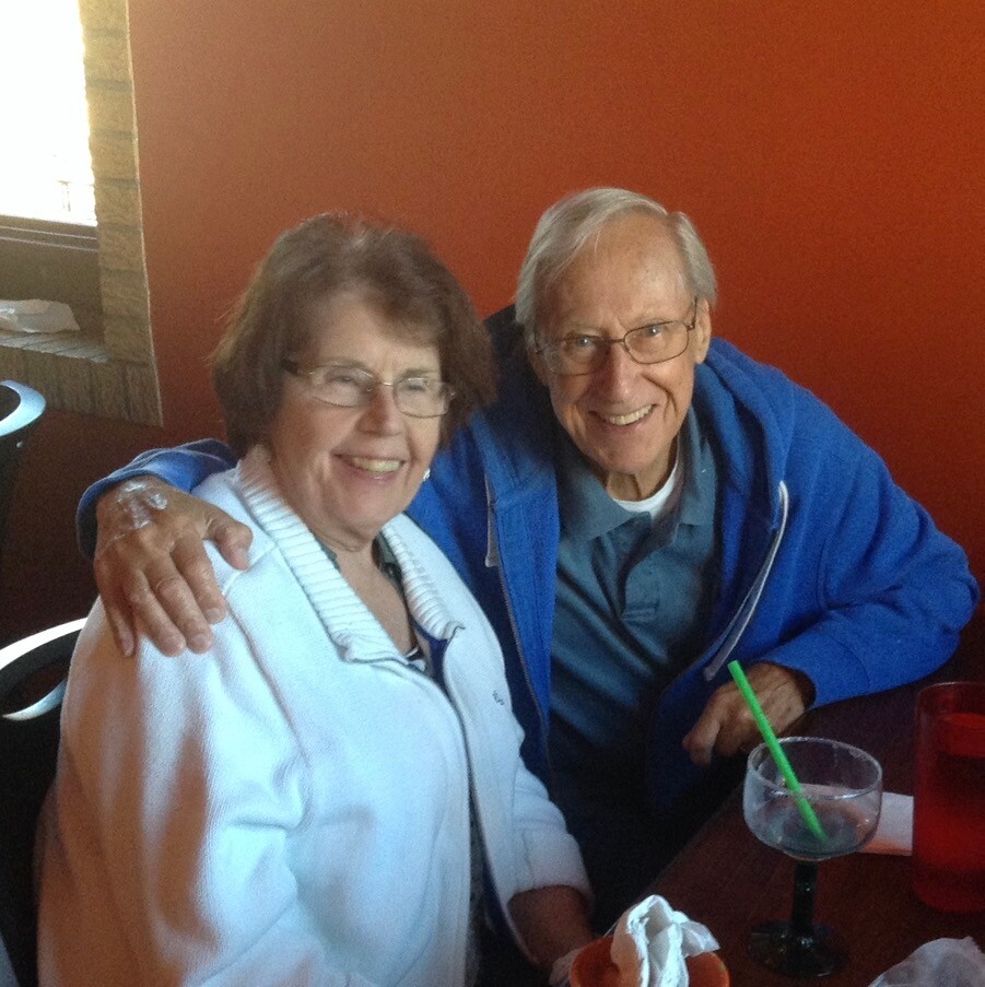 Smiling elderly couple sitting together at a table, with arm around each other, drinks on the table and an orange wall in the background.