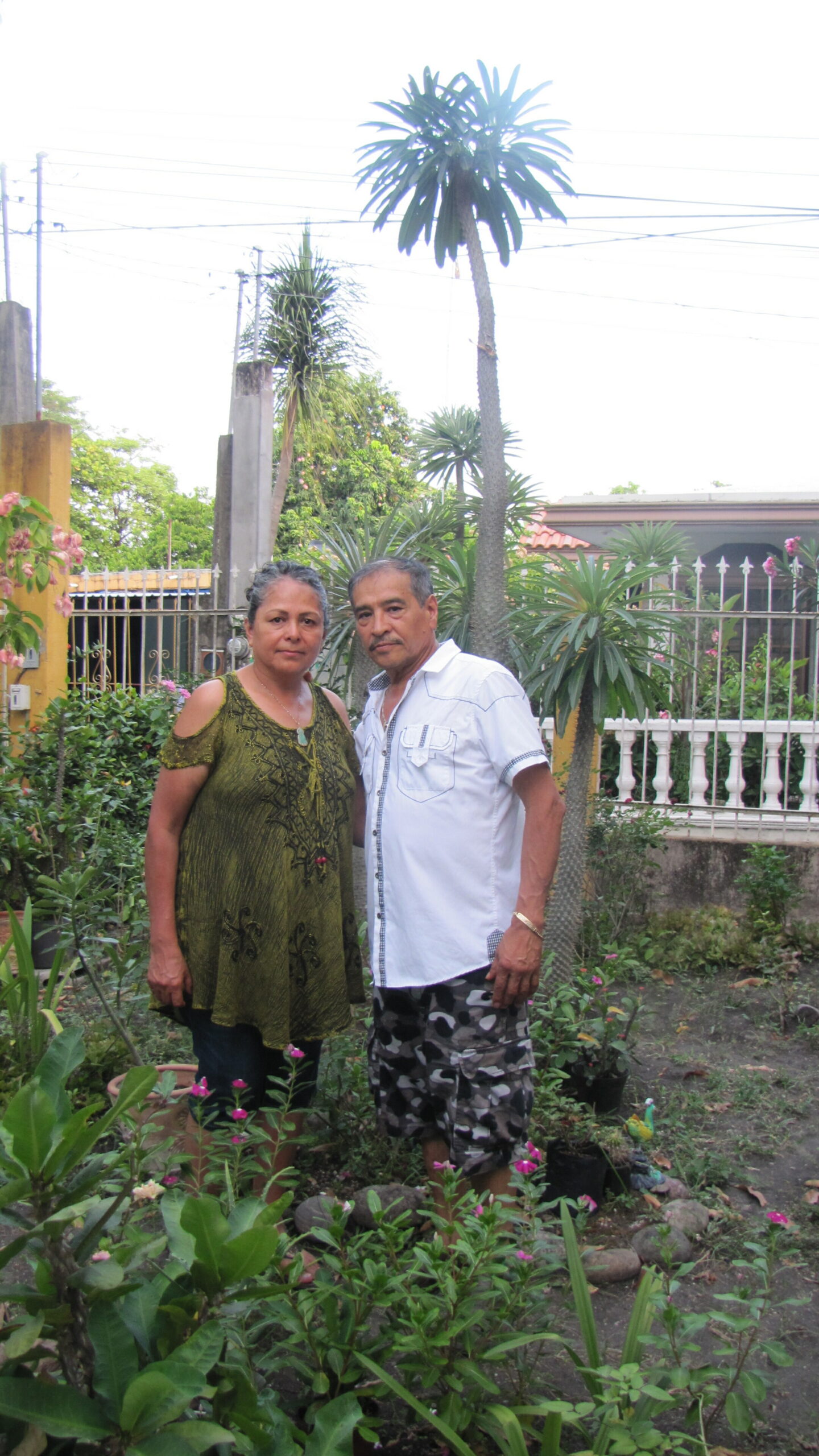 A couple stands together in a garden with tropical plants and a white fence in the background.