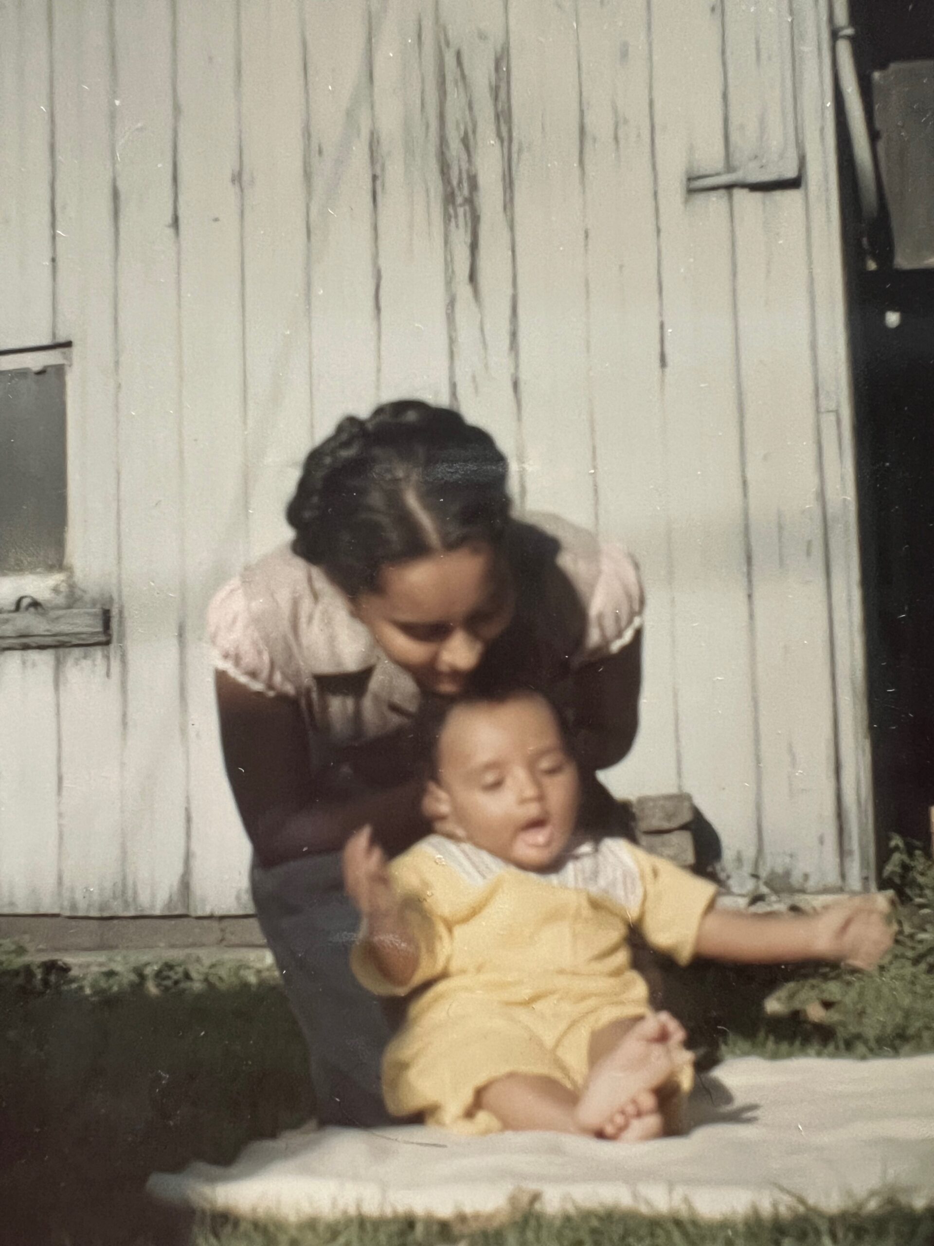 Mother crouching behind a sleeping baby in a yellow outfit on a step outside a white wooden house, sunlight on the yard.