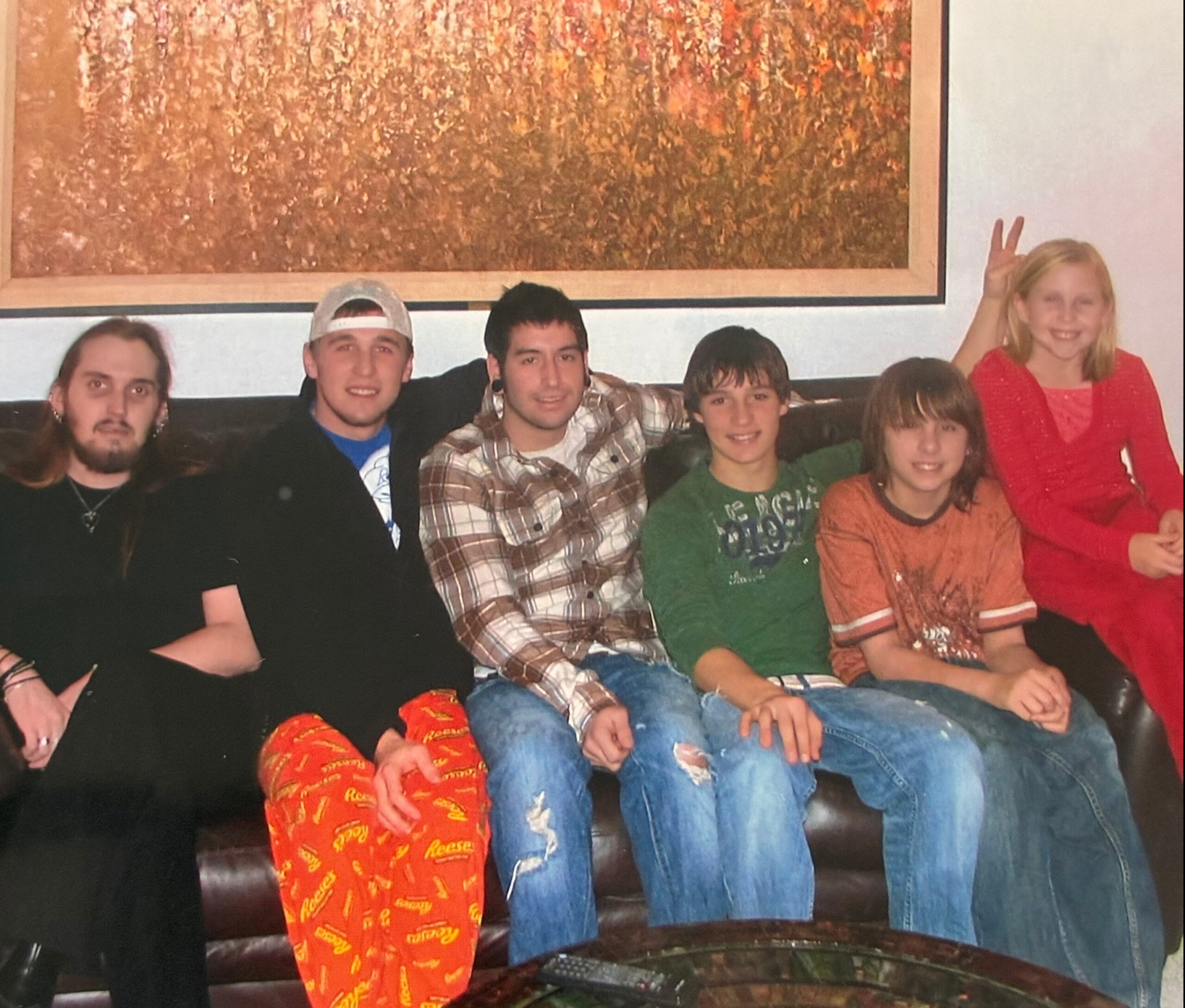 Group of people sitting on a brown leather couch in a living room, posing for a photo with a large abstract painting behind them on the wall.