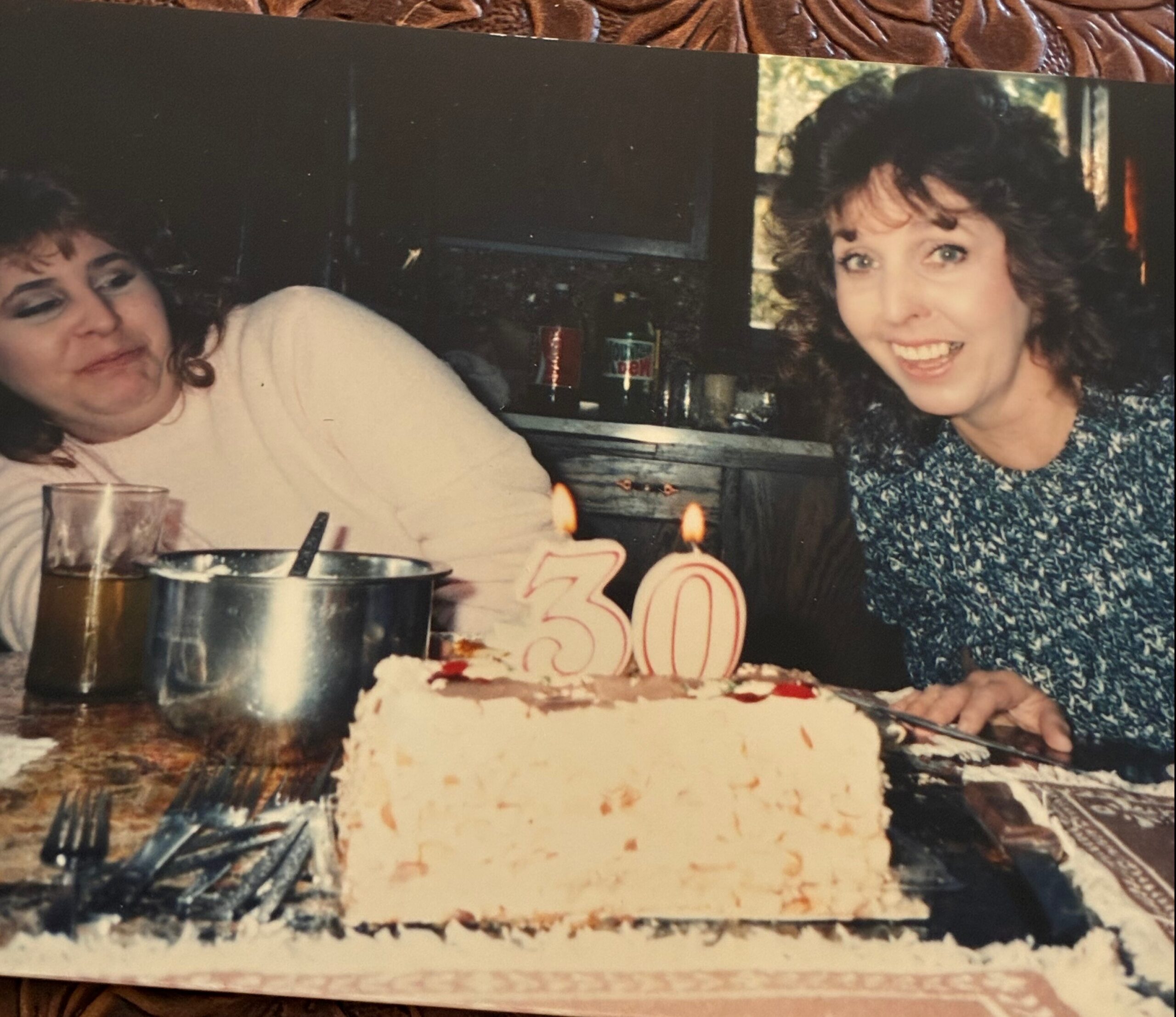 Two women celebrate a birthday at a table with a frosted cake and a drawn '30' candle; one smiles toward the camera while the other looks away.
