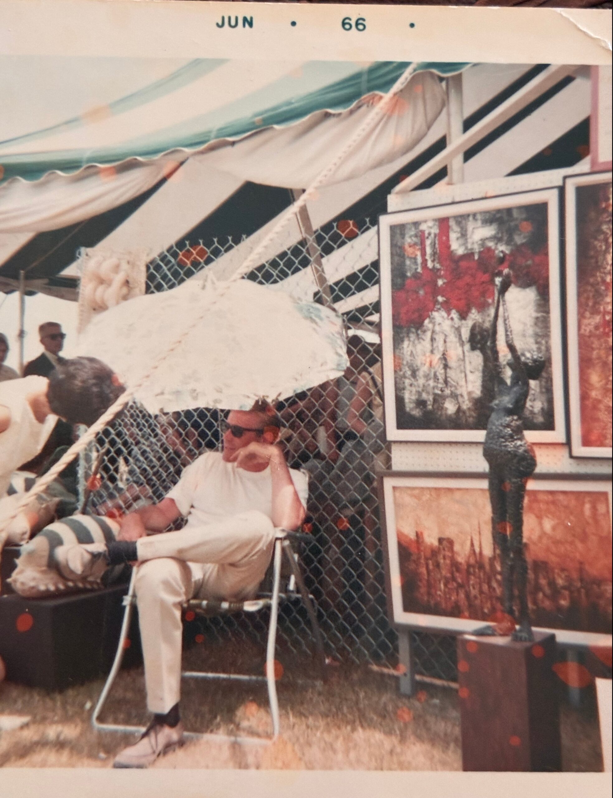 Man in white shirt and light pants seated on a chair at an outdoor art market booth, framed artworks displayed behind a chain-link fence under a striped canopy.