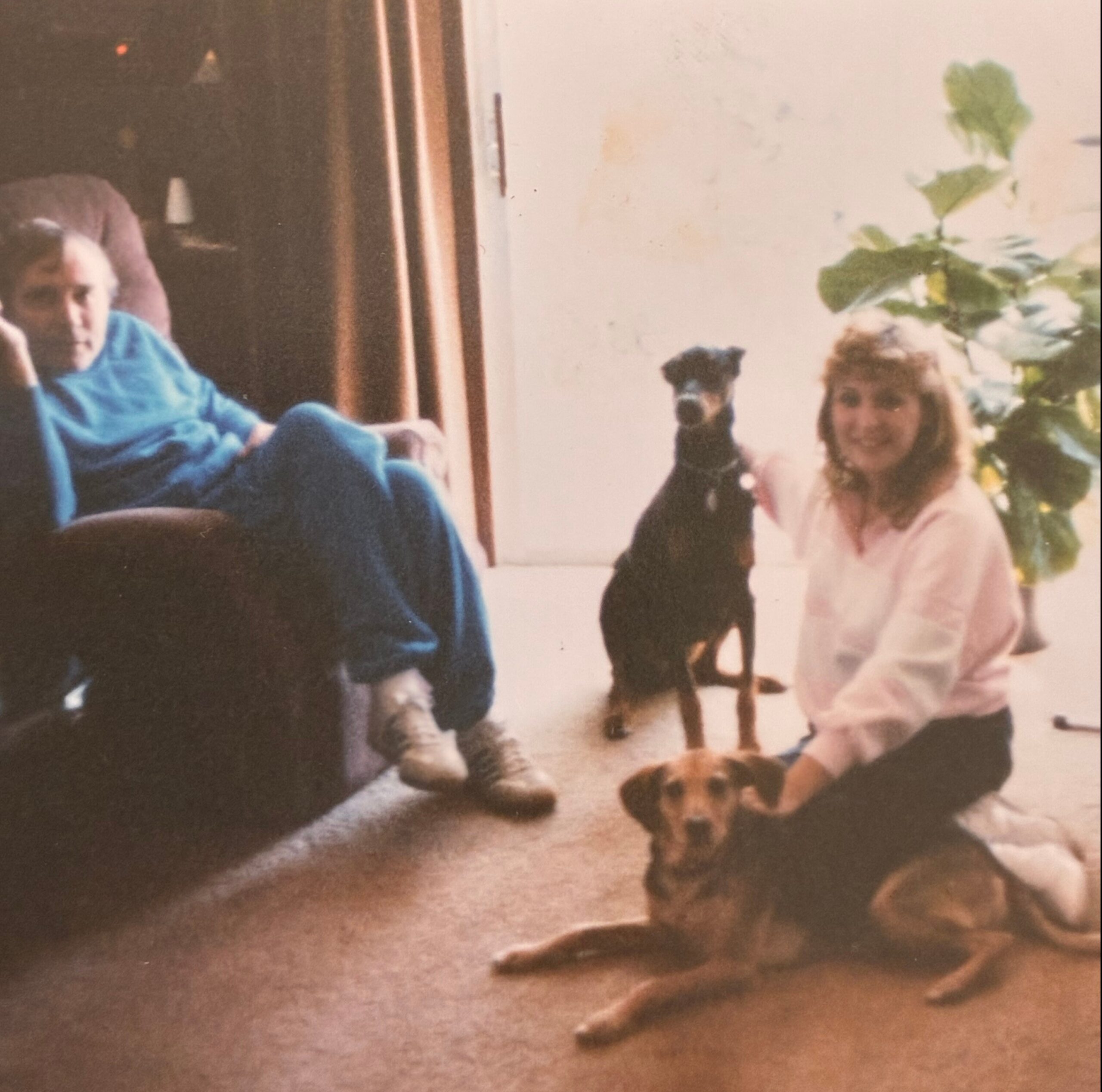 Old faded family photo showing people sitting and standing near a doorway, with a large plant on the right side of the frame and warm indoor lighting behind them.