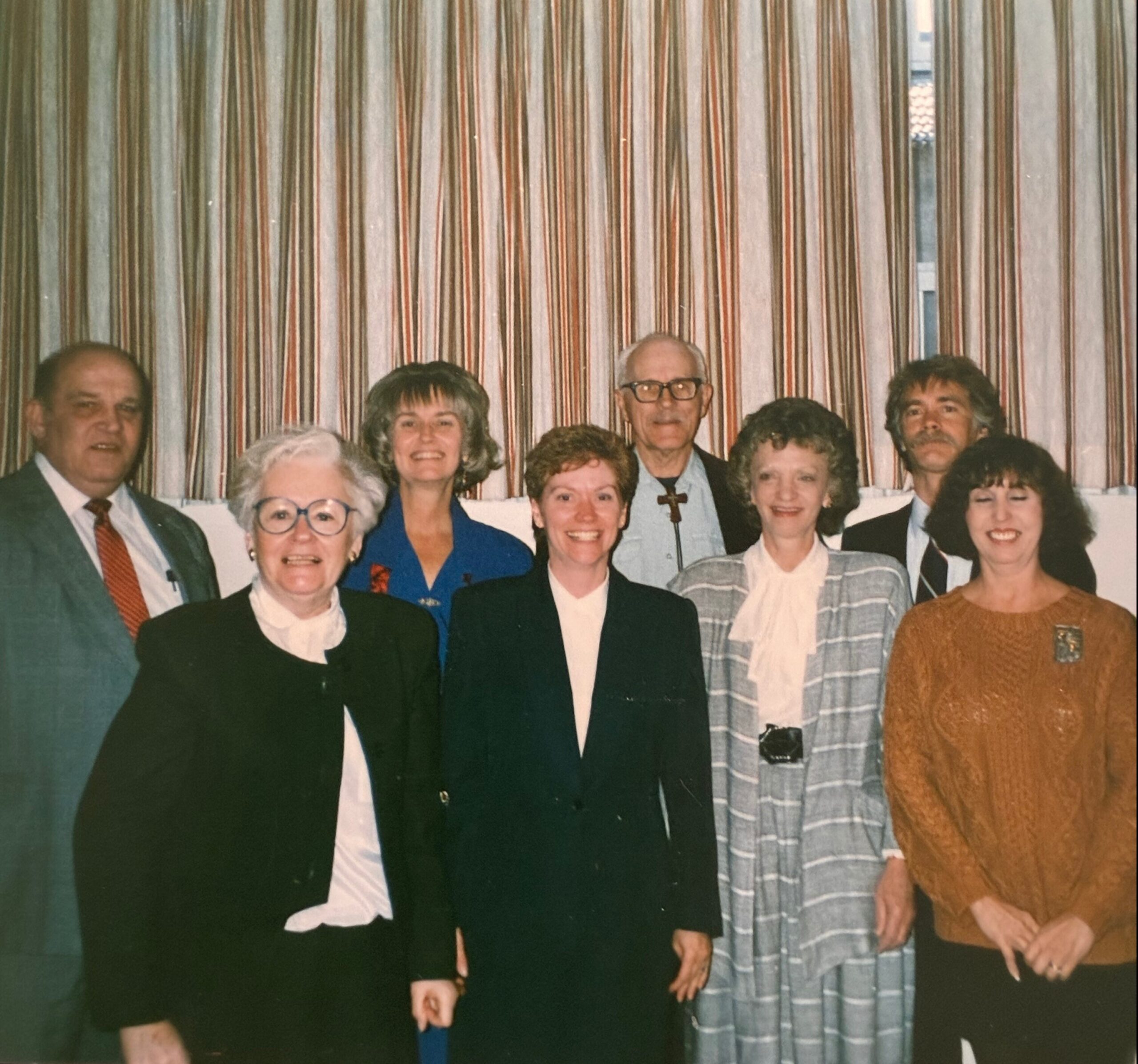 Group of smiling adults posing for a photo indoors, standing in front of striped curtains at a social gathering.