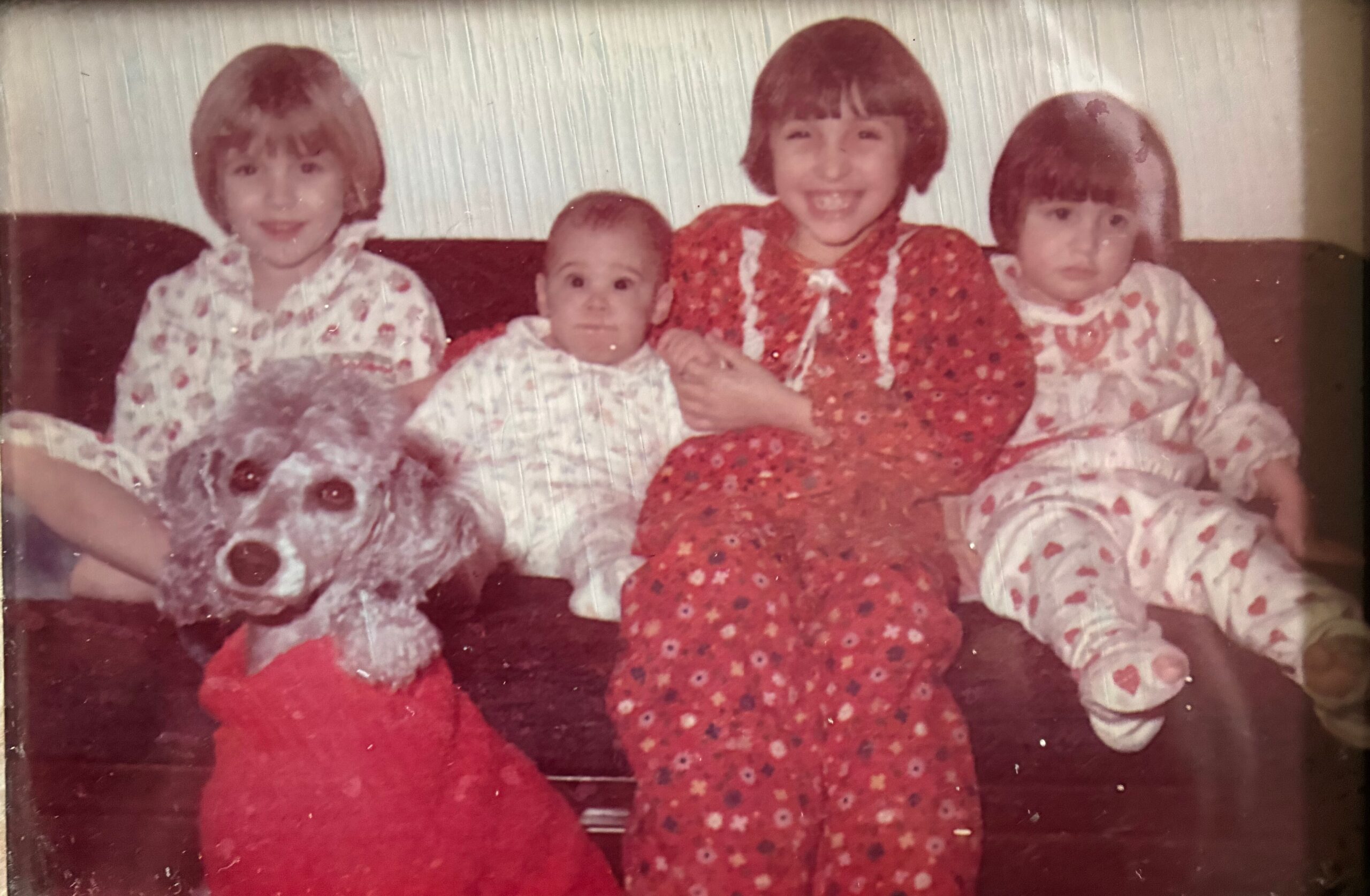 Four children and a baby sit close together on a couch in matching red floral outfits; a fluffy white dog sits at the left.