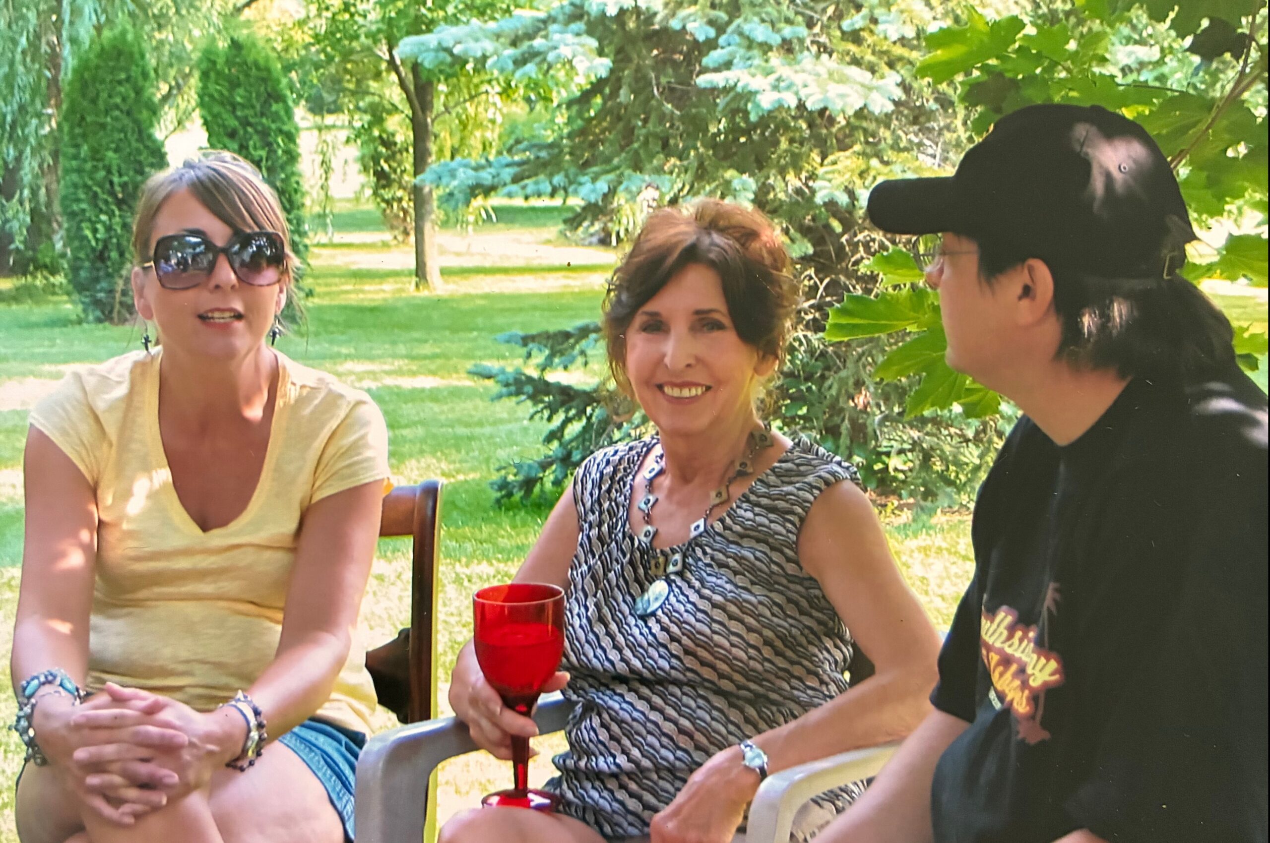 Three adults sit outdoors in a sunny park, chatting, with one holding a red wine glass. The scene shows greenery and casual summer attire.