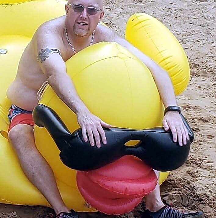 Man wearing sunglasses sits on a large yellow inflatable duck at the beach, shirtless in red swim trunks with a necklace and tattoo visible on his arm.