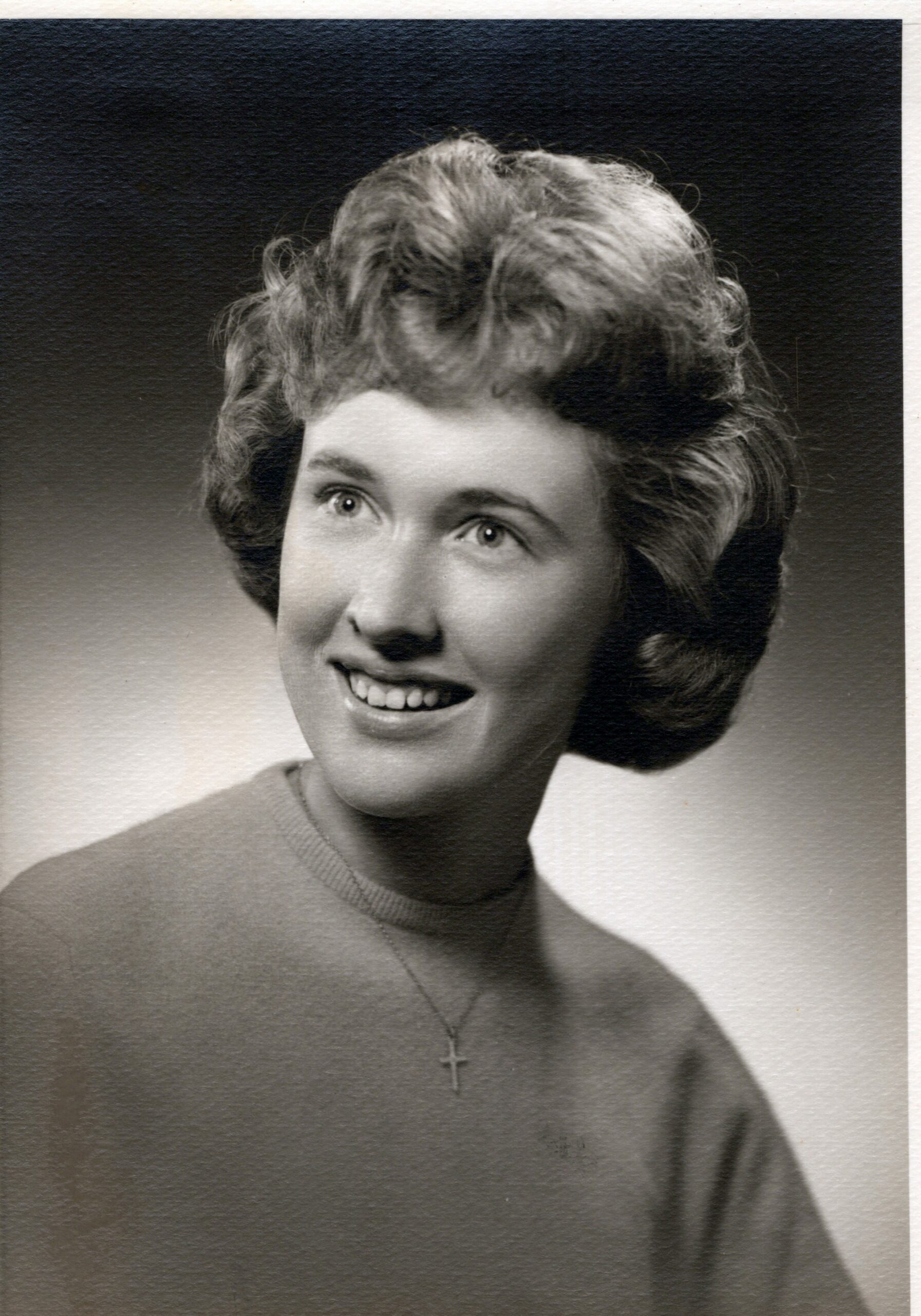Black-and-white portrait of a smiling young woman with short, curled hair, wearing a cross necklace.
