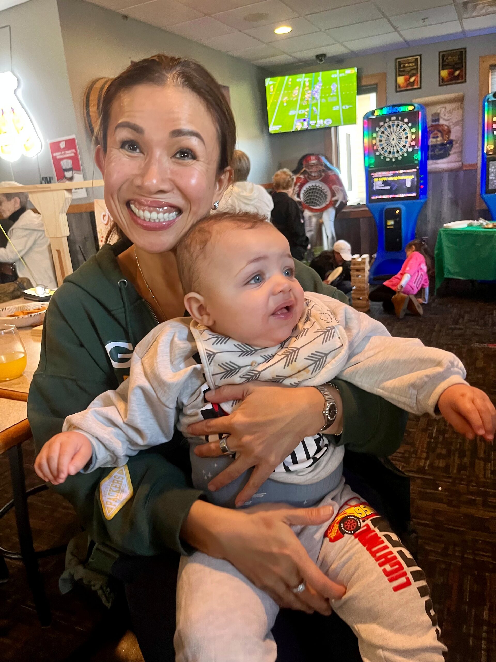 Smiling woman in a green jacket holding a baby in a cafe with arcade games in the background nearby menues and lights behind them.