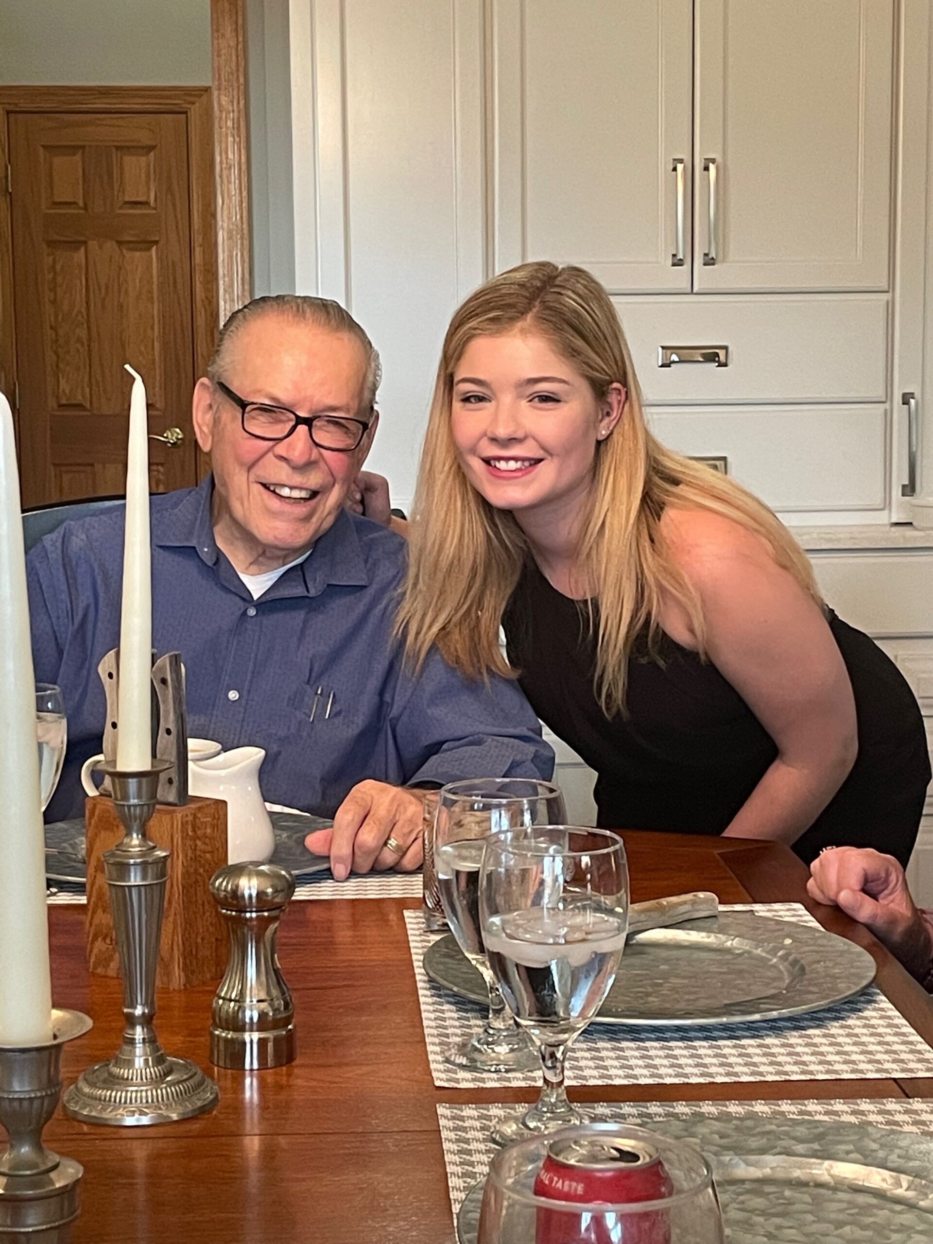 Two people sit at a dining table: an older man with glasses and a blue shirt, smiling beside a younger woman in a black top in a kitchen.