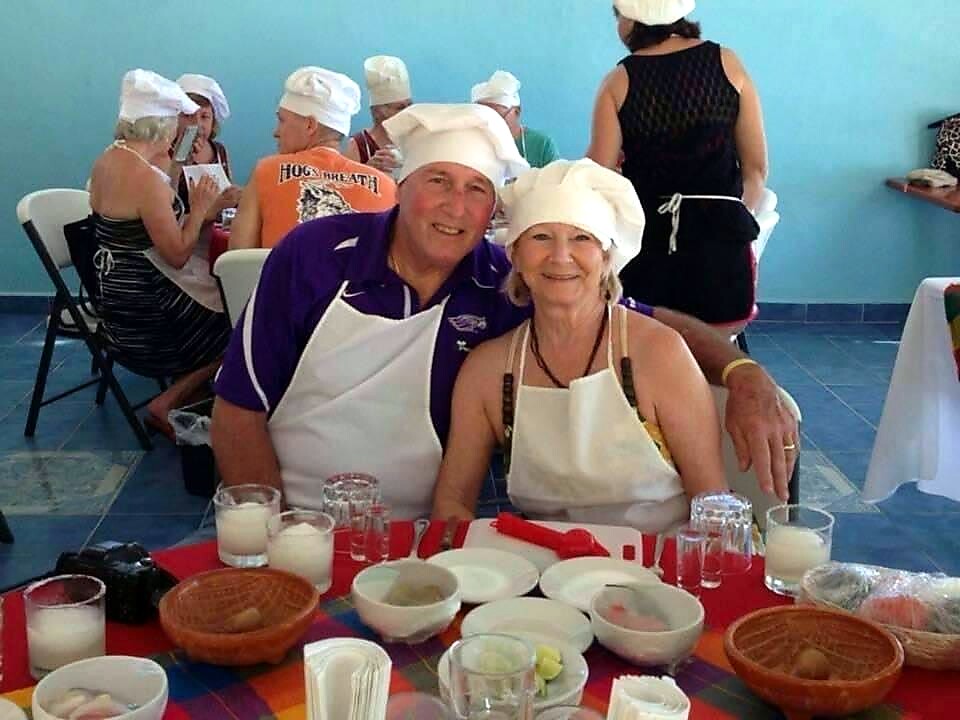 Smiling couple wearing chef hats and aprons sit at a dining table during a cooking class, with other participants in the background.