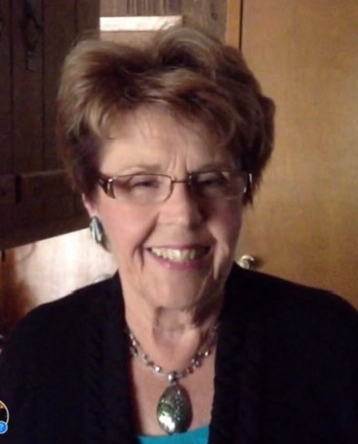 Smiling older woman with short brown hair and glasses, wearing a turquoise top and green pendant necklace indoors.