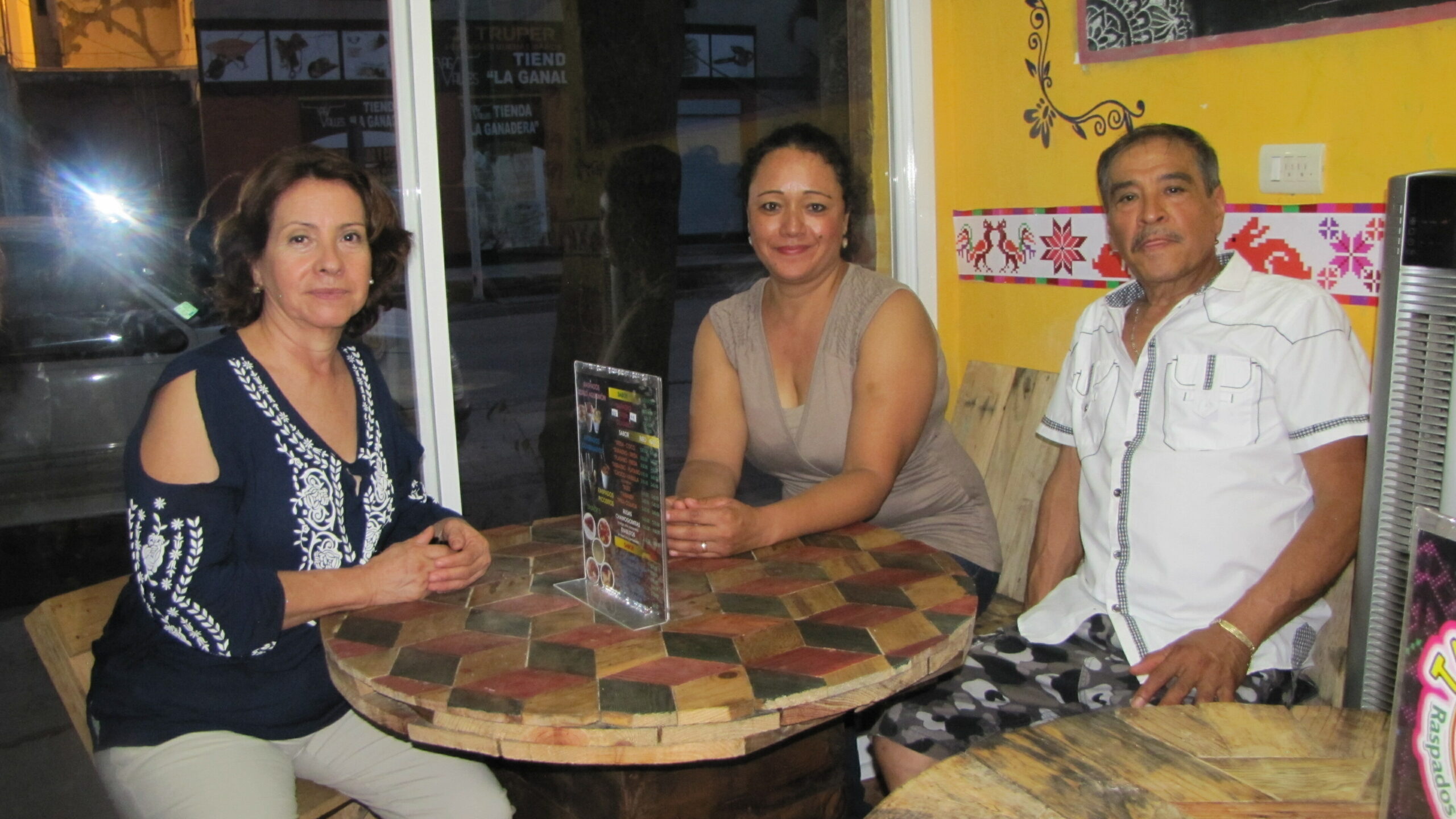 Three people sit at a rustic, multi-toned wooden table in a cafe, posing for the camera with smiles. A woman in blue with embroidery on the left, a woman in beige in the middle, and a man in a white shirt on the right, colorful wall art behind them.