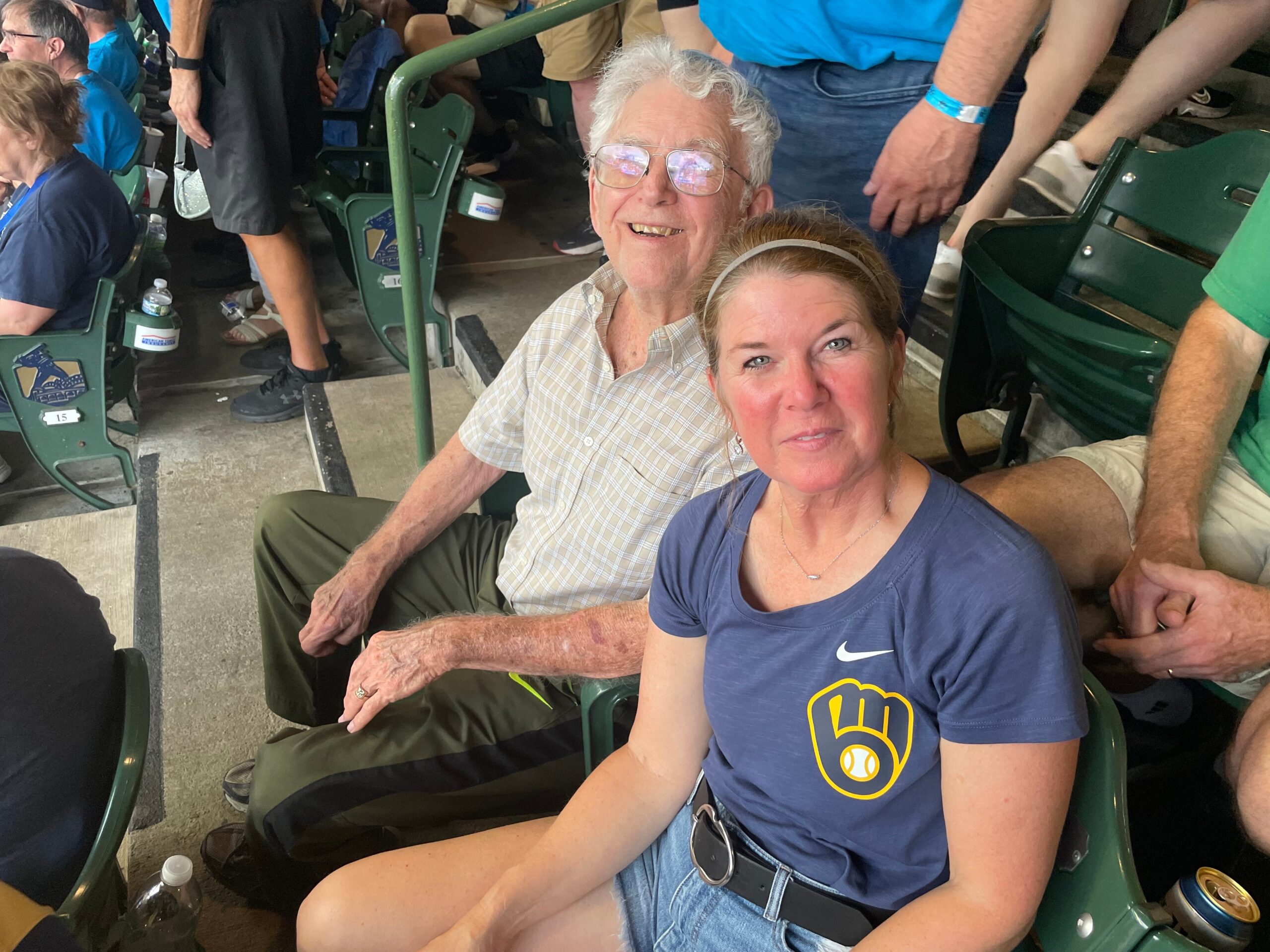Smiling elderly man with white hair and glasses seated in a stadium beside a woman in a blue shirt, both looking at the camera amid other spectators and green seats.