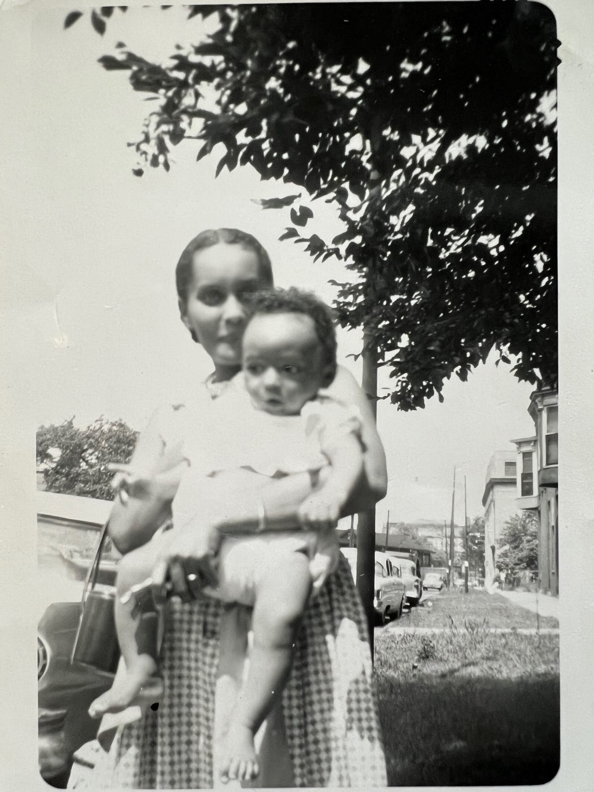 Two young children standing outdoors on a street, one holding the other, with a tree and houses in the background.