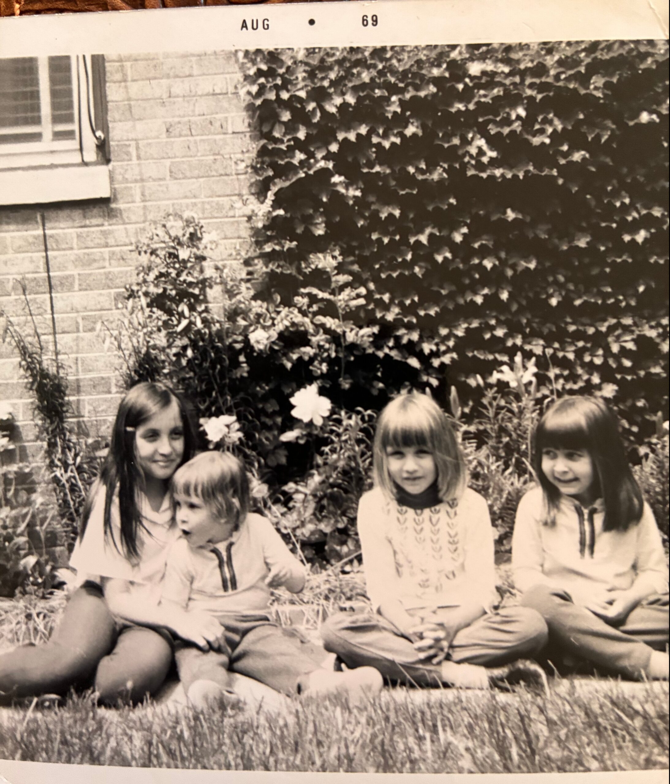 Group of young children sitting on the grass in front of a brick house covered with ivy and flowers, posing for a photo (1969).