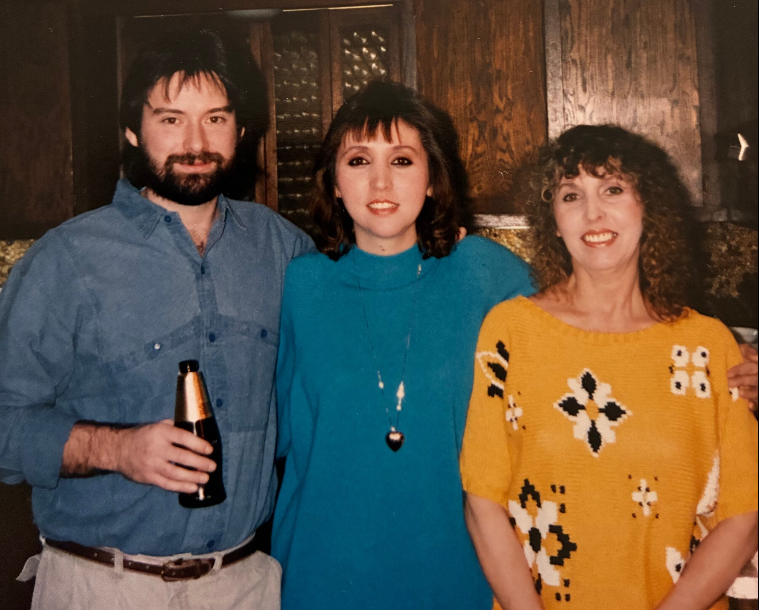 Three adults posing for a casual photo indoors: man in blue shirt holding a beer bottle on the left, a woman in teal in the middle, and a woman in a yellow owl-pattern sweater on the right.