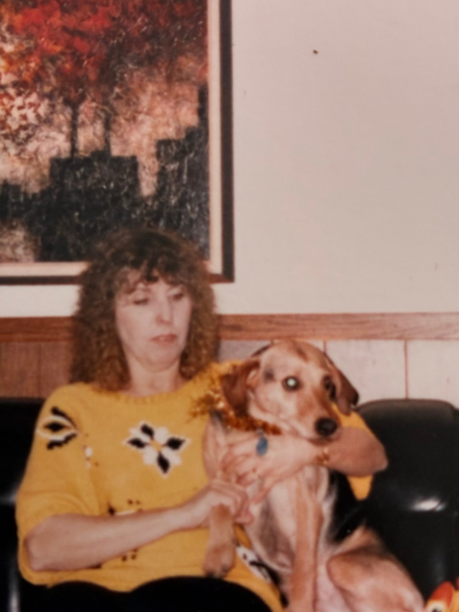 Woman with curly hair in a yellow sweater holding a small dog on their lap indoors, with a painting in the background.
