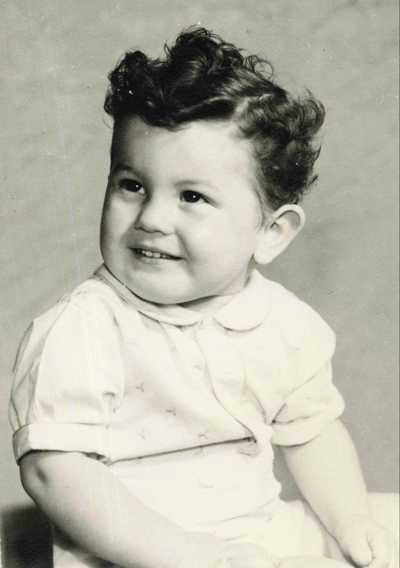 Smiling baby with dark, curly hair wearing a light shirt, looking to the side.