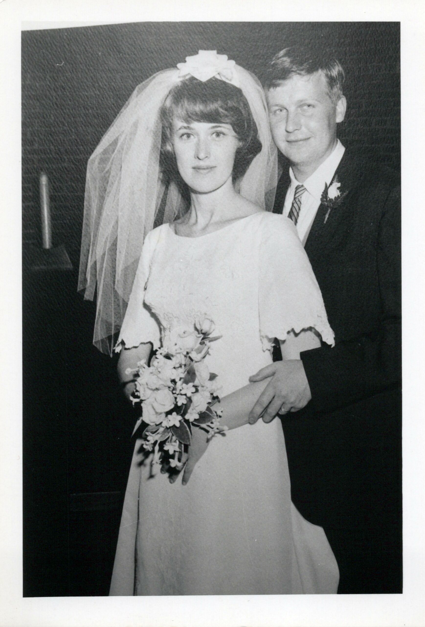 Bride in a veil and wedding dress holding a bouquet, standing with a groom in a dark suit at their wedding.