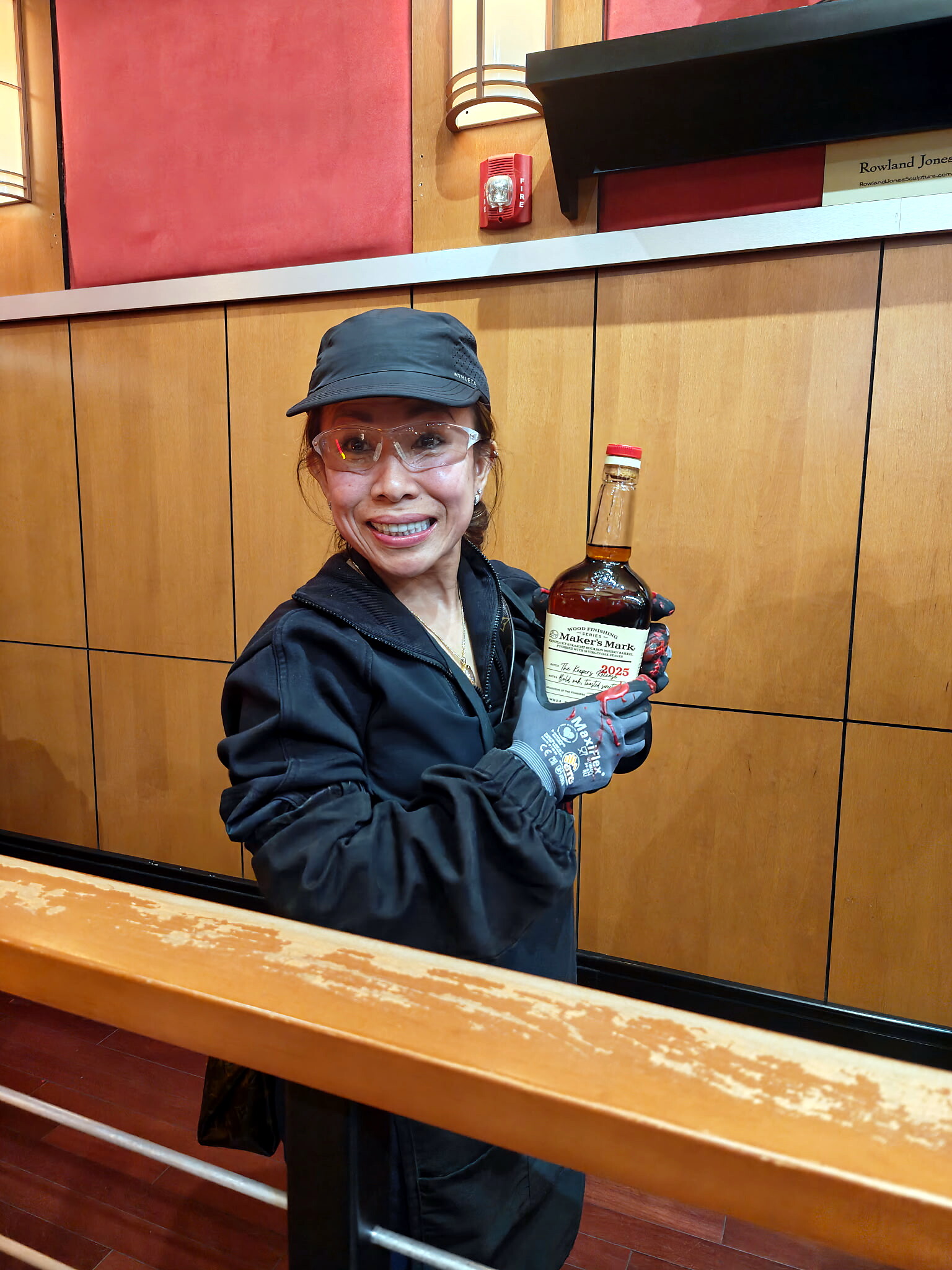Smiling woman in a cap and safety glasses holds a Maker's Mark bourbon bottle in a wood-paneled room, likely a bar.