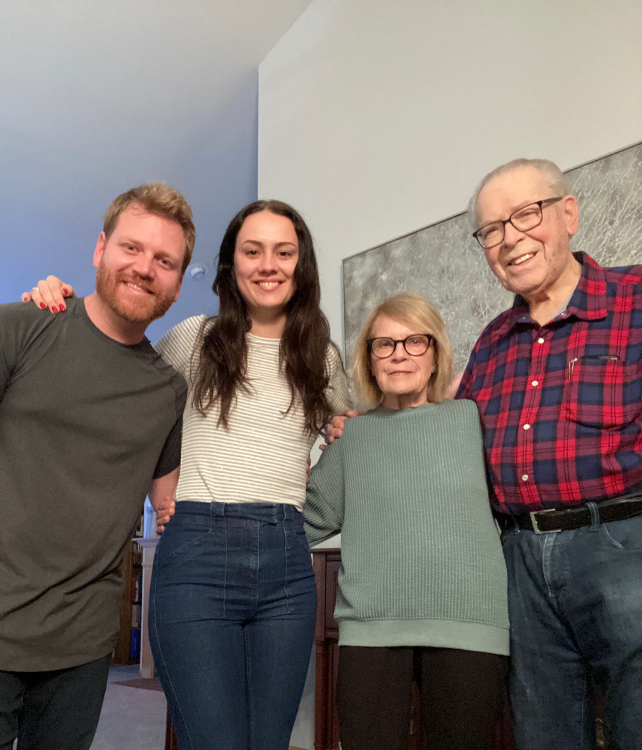 Four adults posing together indoors, smiling at the camera; left man in olive shirt, woman in striped top, older woman with blond hair wearing a green sweater, and man in a red plaid shirt on the right, with a blue wall and furniture in the background.