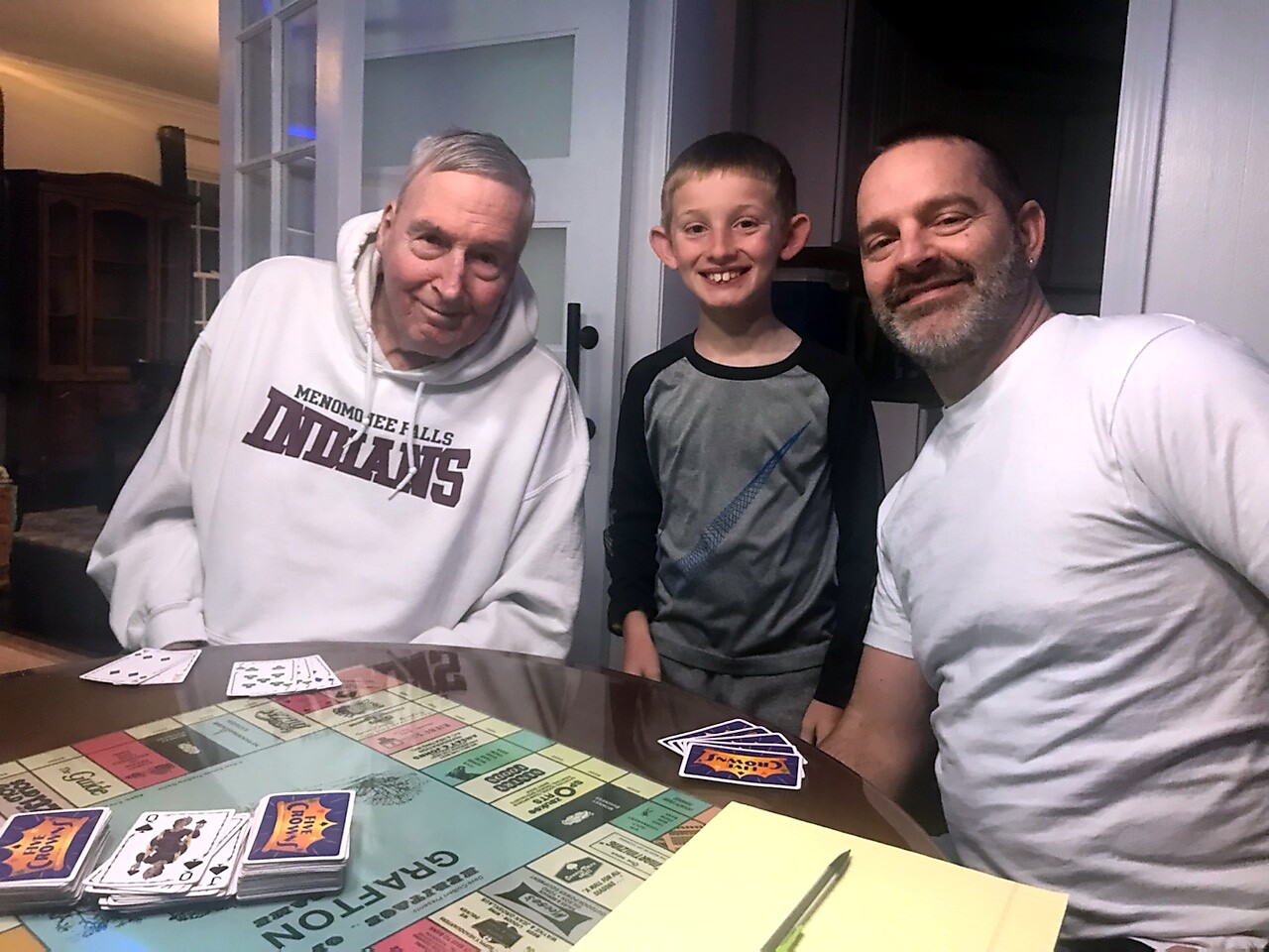 Three smiling people sit around a colorful board game on a kitchen table with cards laid out nearby, in a home setting.