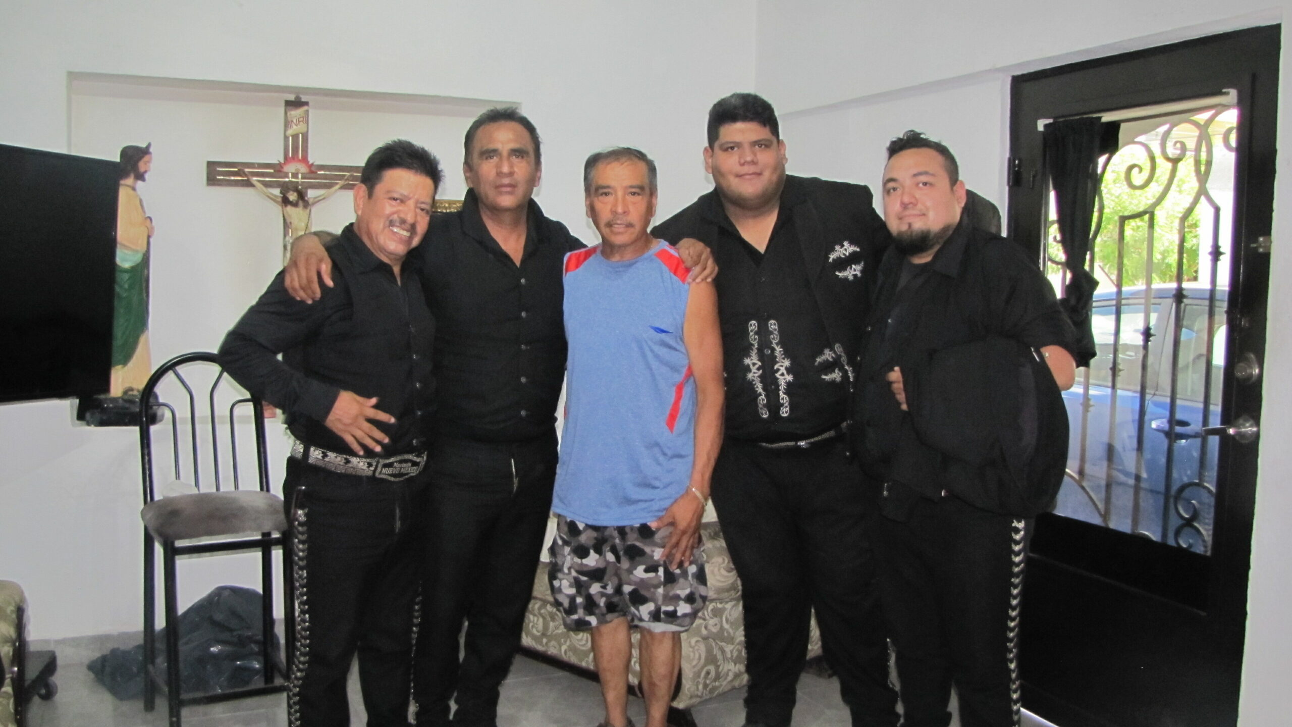 Group of five men standing together indoors, one in a blue athletic shirt and camo shorts flanked by four men in black outfits.