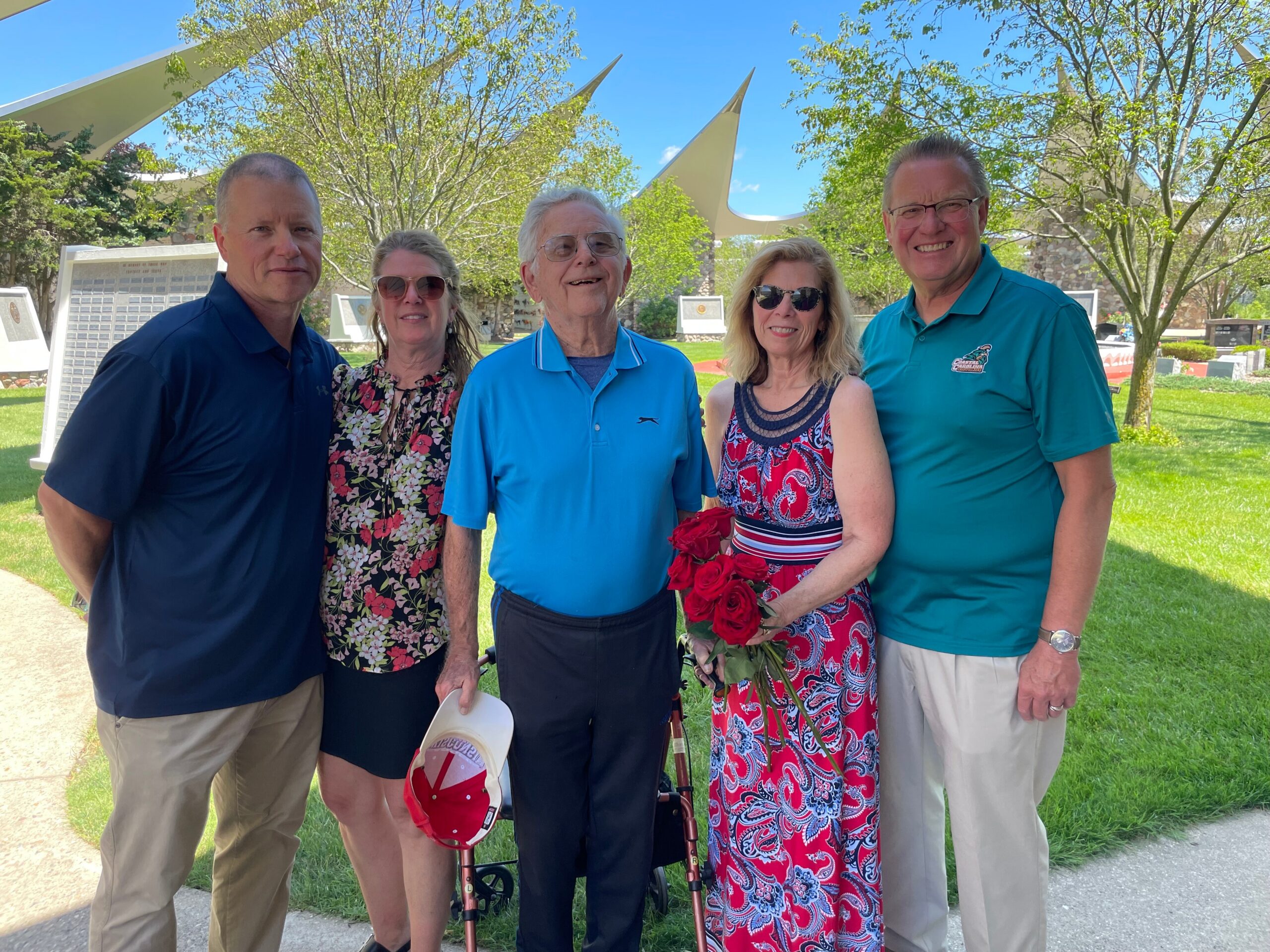 Five adults posing together outdoors on a sunny day, smiling with trees and a sculpture in the background.