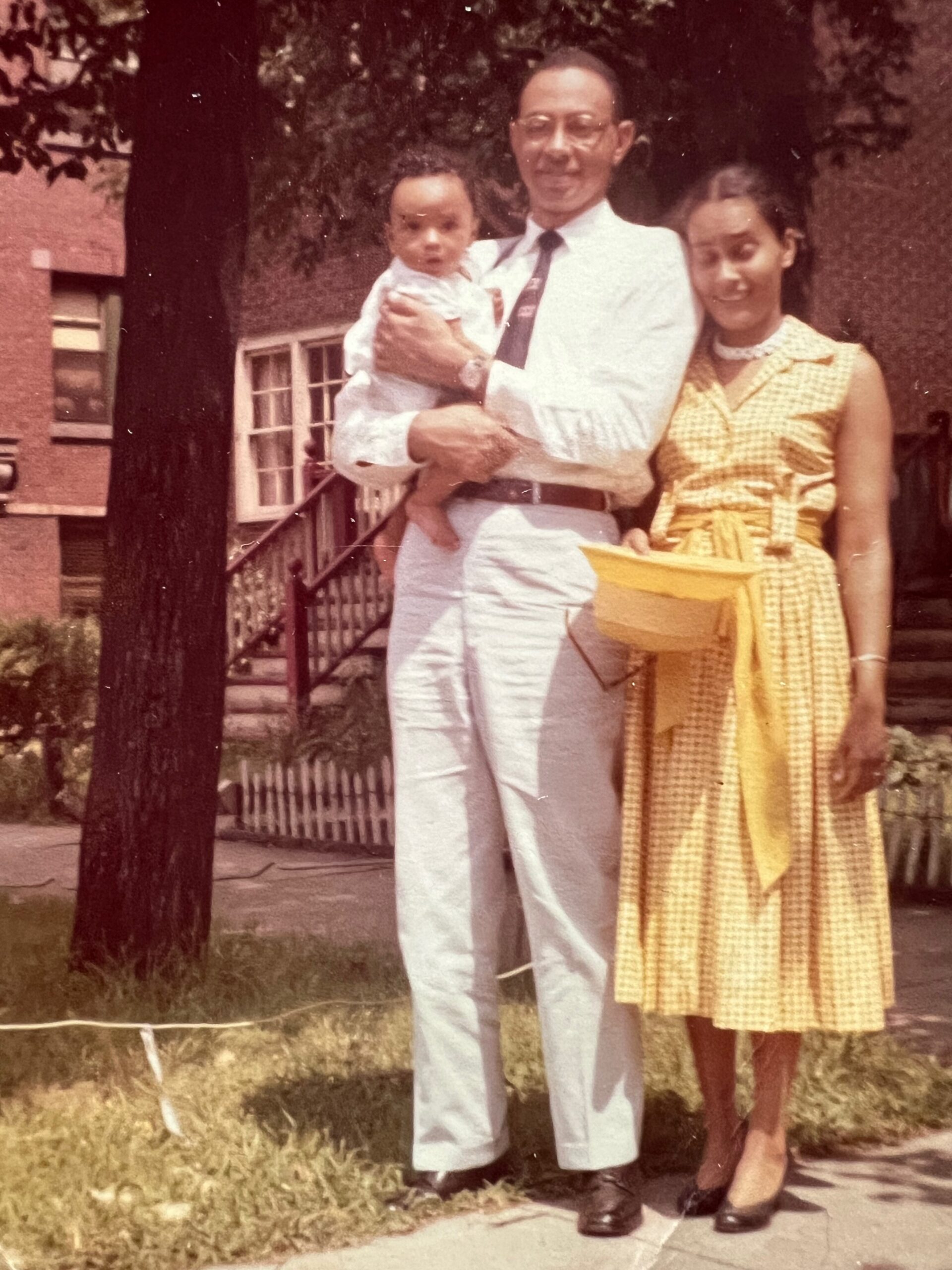 Father in a white suit holds a baby while his wife in a yellow gingham dress stands beside them outdoors in a residential yard with a brick building nearby.