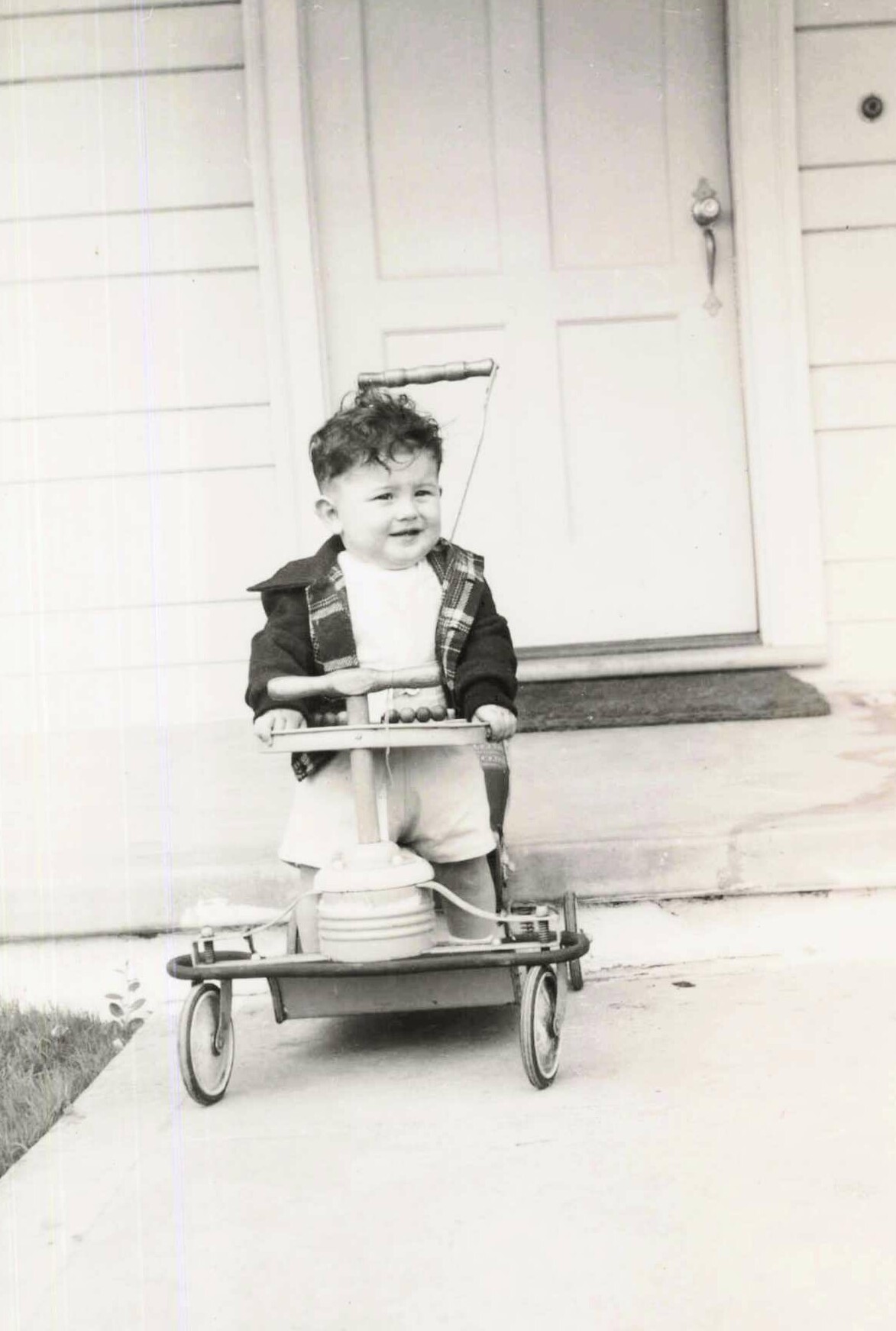 Toddler on a vintage tricycle in front of a house door, wearing a plaid jacket and white outfit.