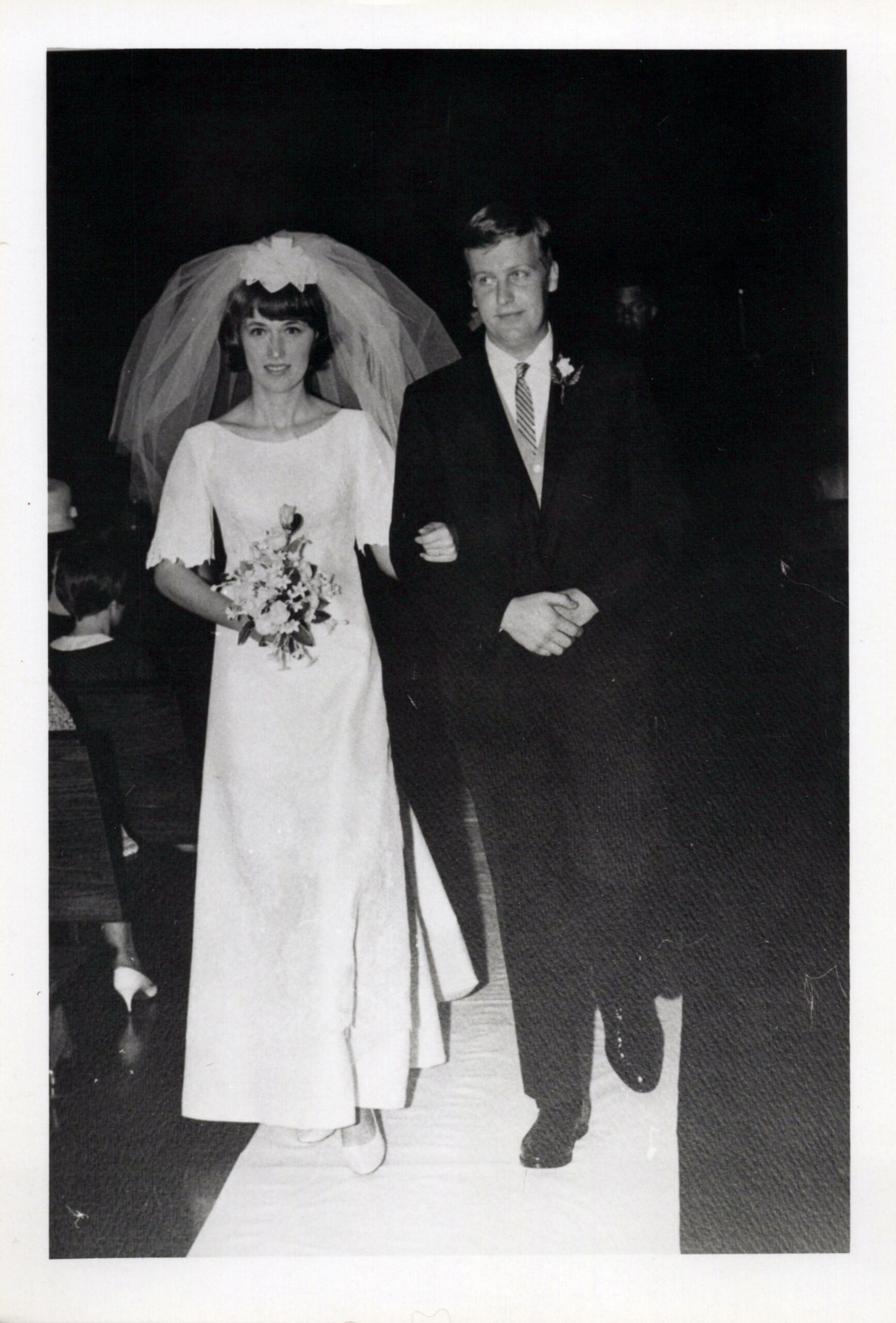 Bride in a white wedding dress with a veil holding a bouquet, walking arm-in-arm with a groom in a dark suit and striped tie.