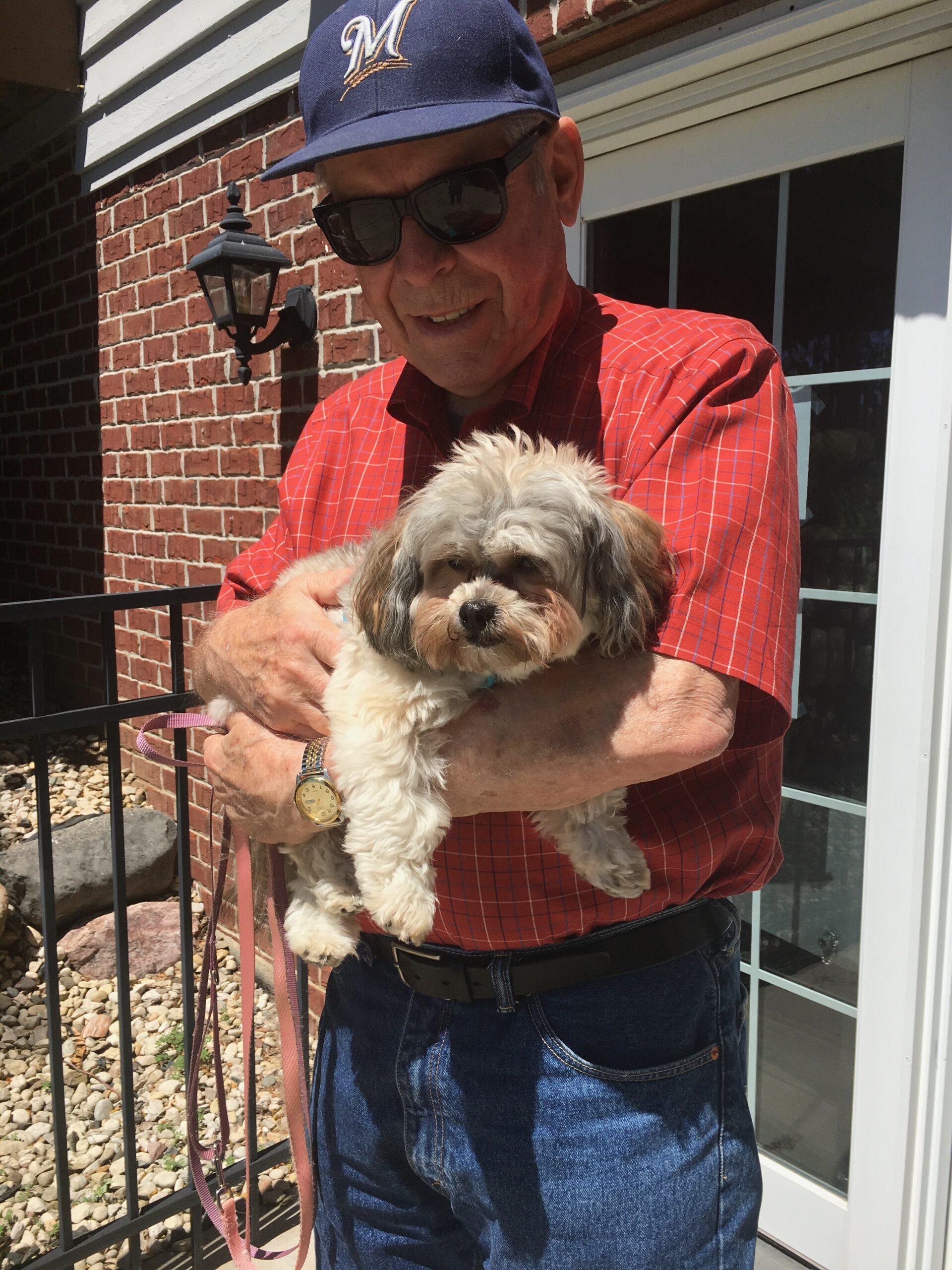 Older man wearing a blue cap and sunglasses holds a small fluffy dog outdoors beside a brick house.