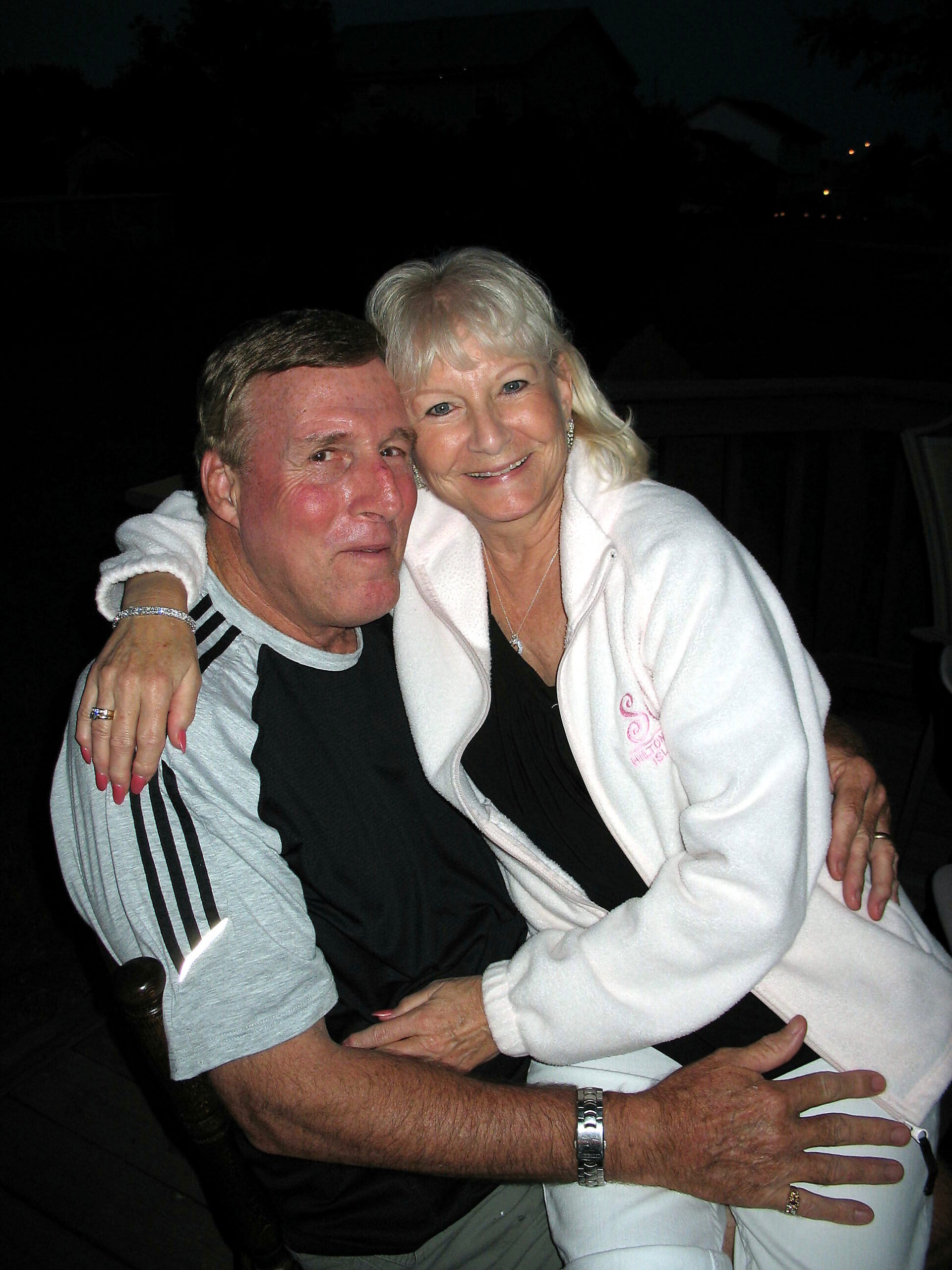 Smiling elderly couple embracing each other outdoors at night, arms around one another.