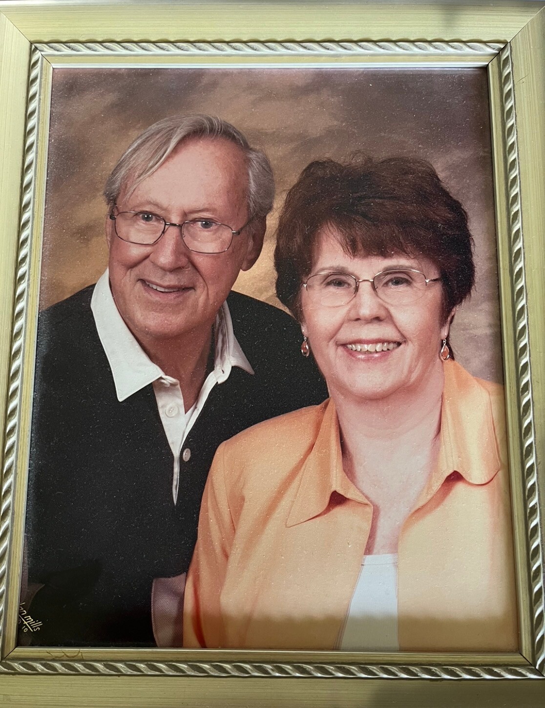 Framed portrait of an elderly couple smiling; man with glasses in a dark sweater and white shirt, woman in an orange blouse wearing glasses.