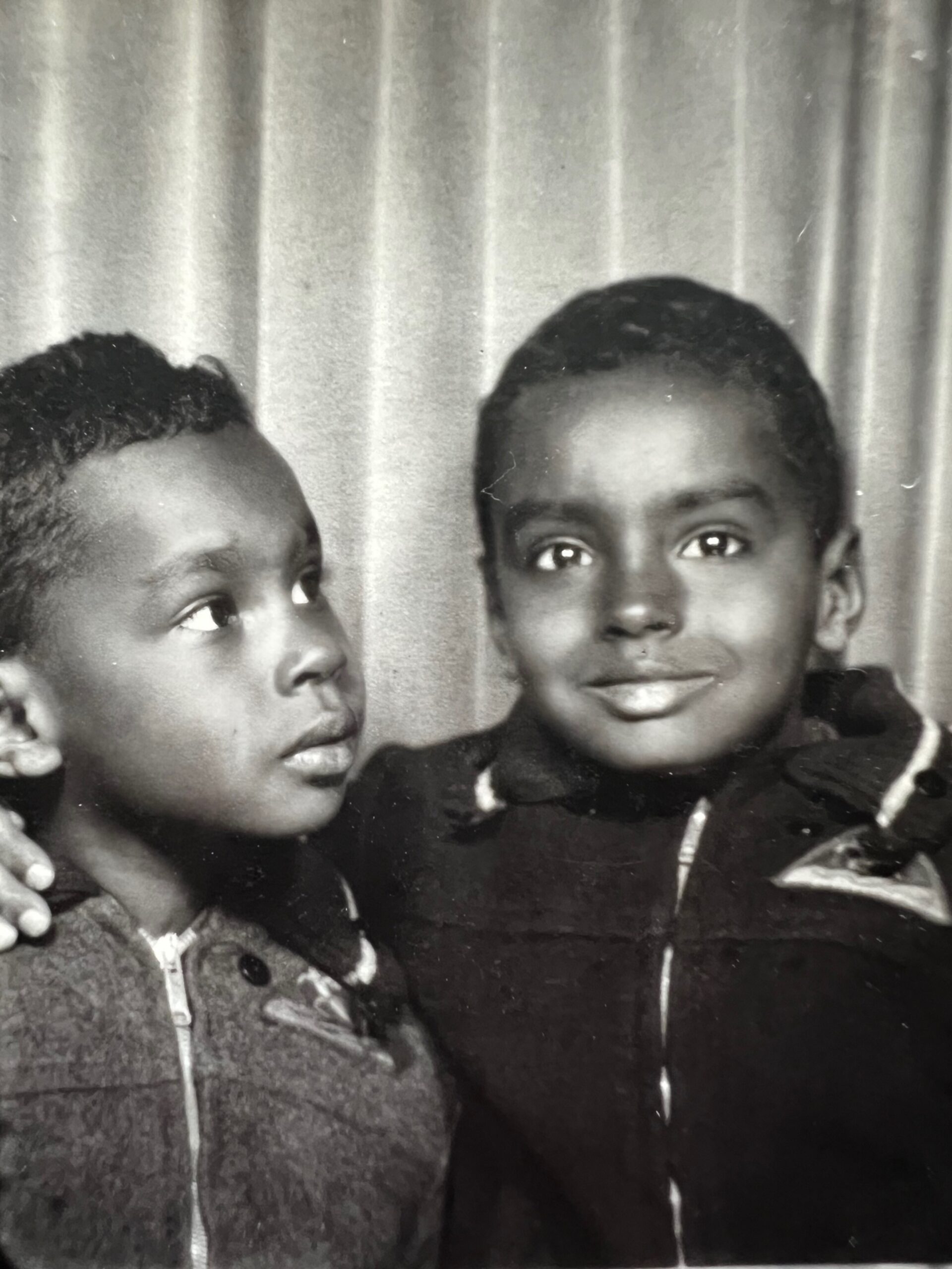 Two young boys in dark jackets posing together in front of a curtain backdrop.