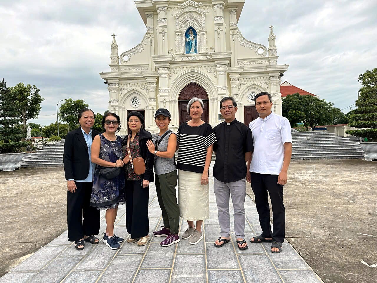 Group of eight adults posing in front of an ornate white church with a statue niche above the entrance; sunny day, casual attire and smiles across the lineupside-by-side.