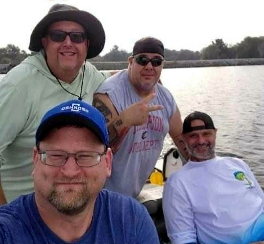 Four men wearing hats and sunglasses pose for a selfie on a boat by a lake, smiling at the camera.