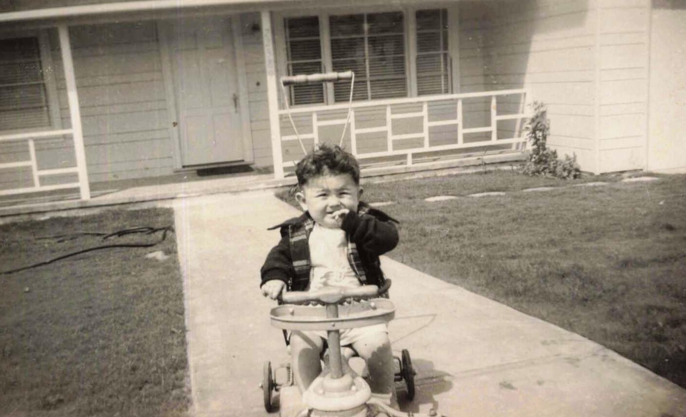Young child on a tricycle riding along a concrete sidewalk in front of a suburban house with a porch.