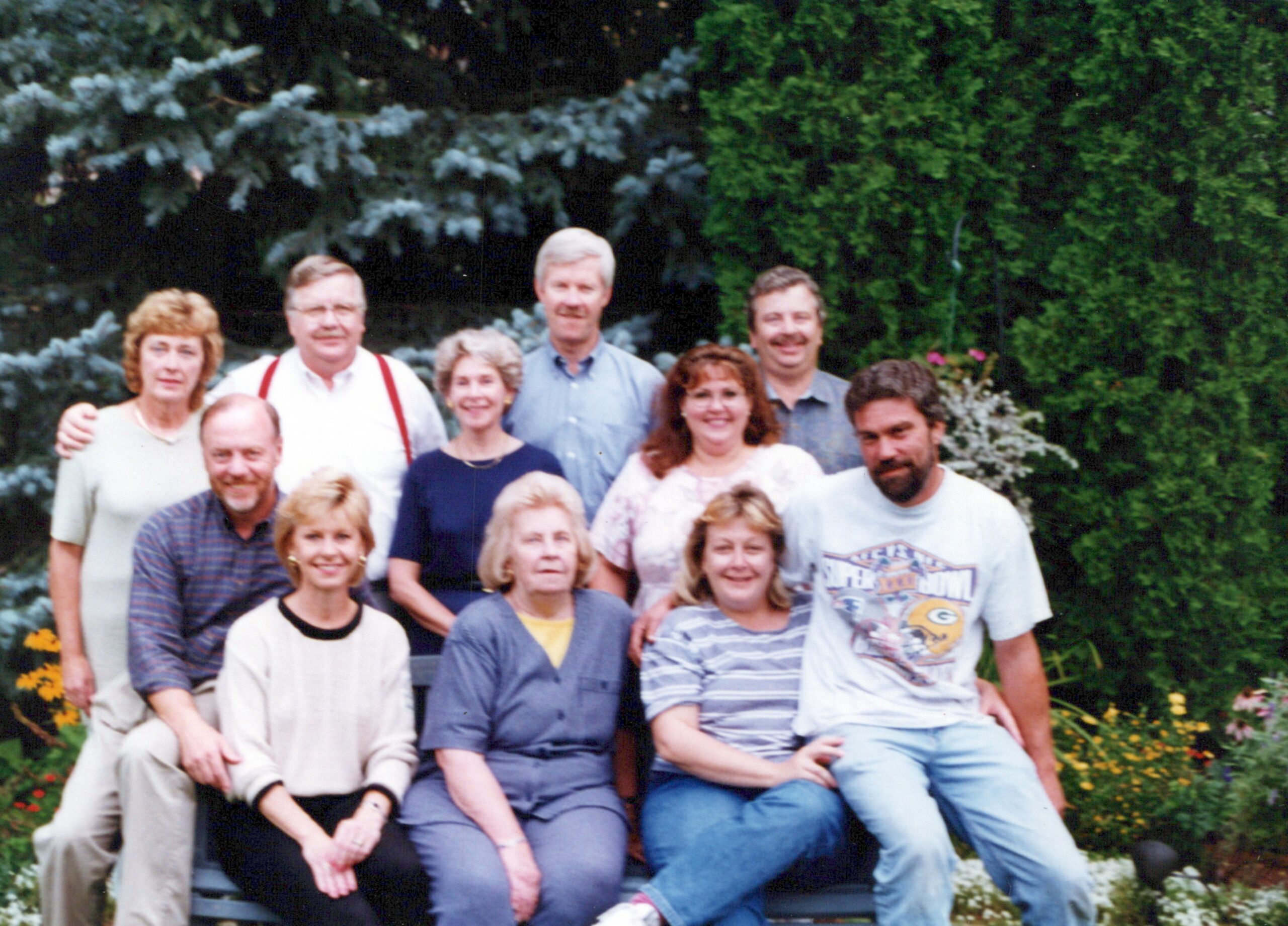 Group of about ten adults posing together outdoors in a garden with green hedges and flowers, some standing and some sitting on a bench outside.