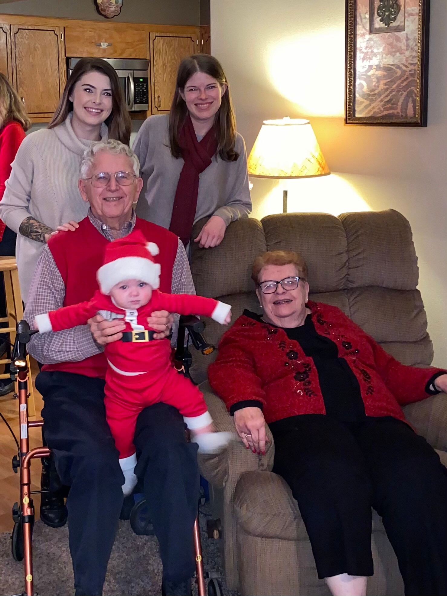 Family photo in a living room: an older man in a red vest holding a baby in a Santa outfit, with two smiling women behind and an elderly woman seated nearby.