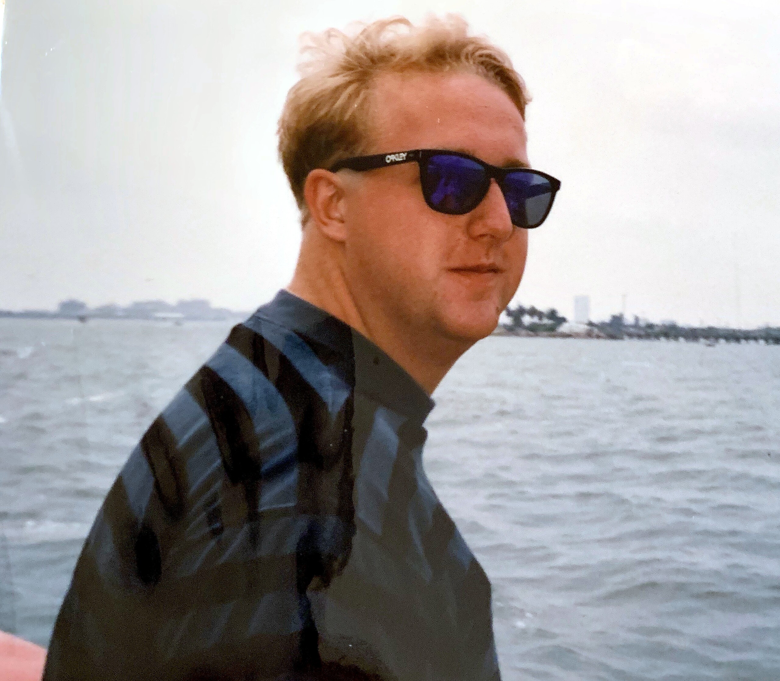 Man wearing dark sunglasses near a body of water, cropped at chest level, with a light sky behind him.