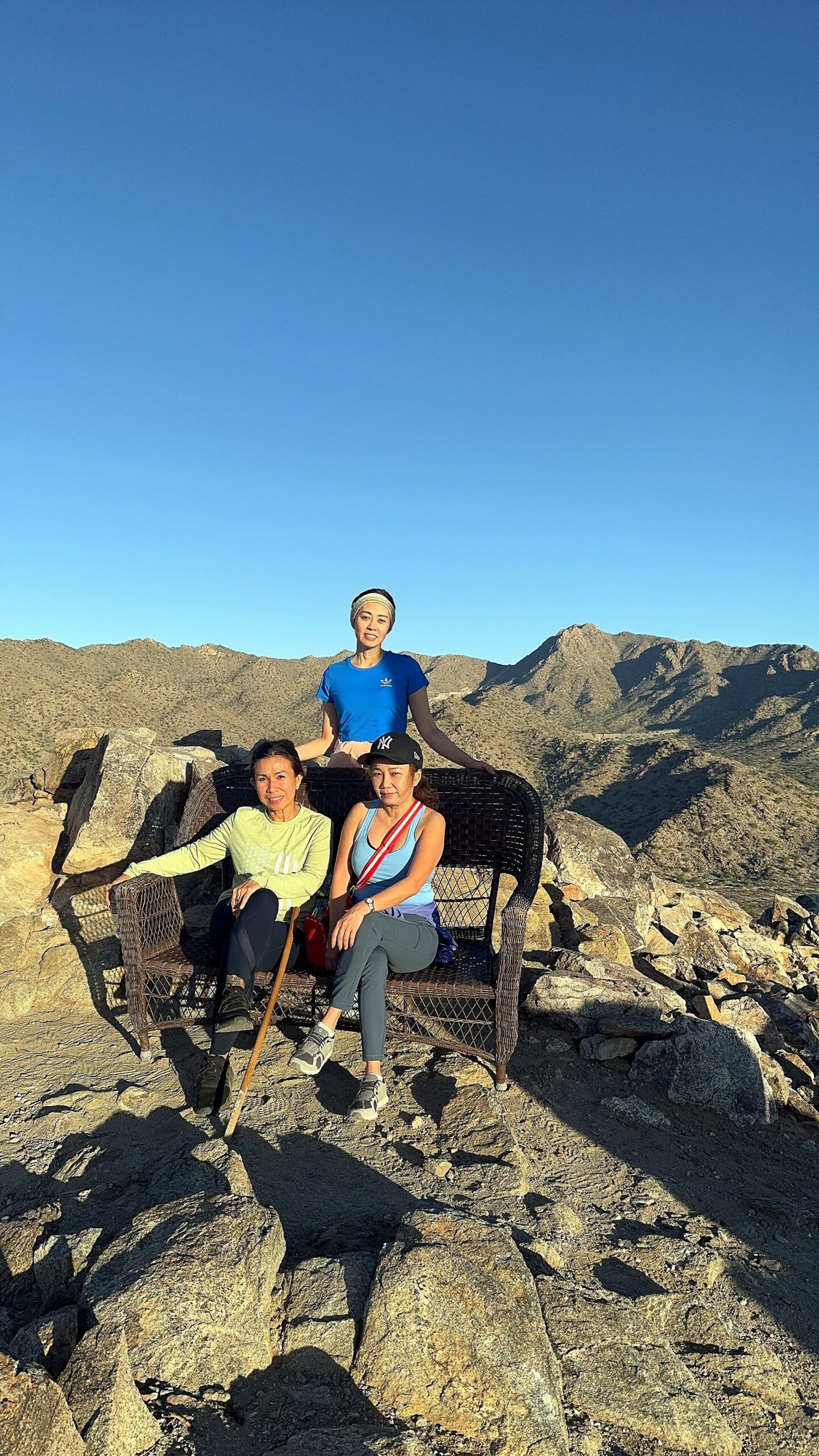 Three women sit on a wicker bench on a rocky mountain summit under a clear blue sky, wearing athletic clothing and caps.
