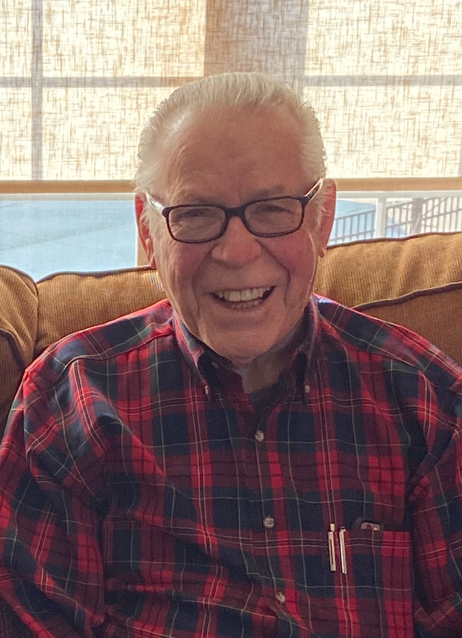 Smiling elderly man with white hair and glasses wearing a red-and-blue plaid shirt, seated indoors on a sofa.