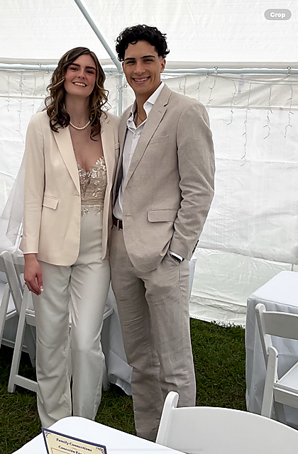 A smiling couple in light beige suits posing together at an outdoor tent event, the woman in a cream blazer and lace top, the man in a matching beige suit with an white shirt.