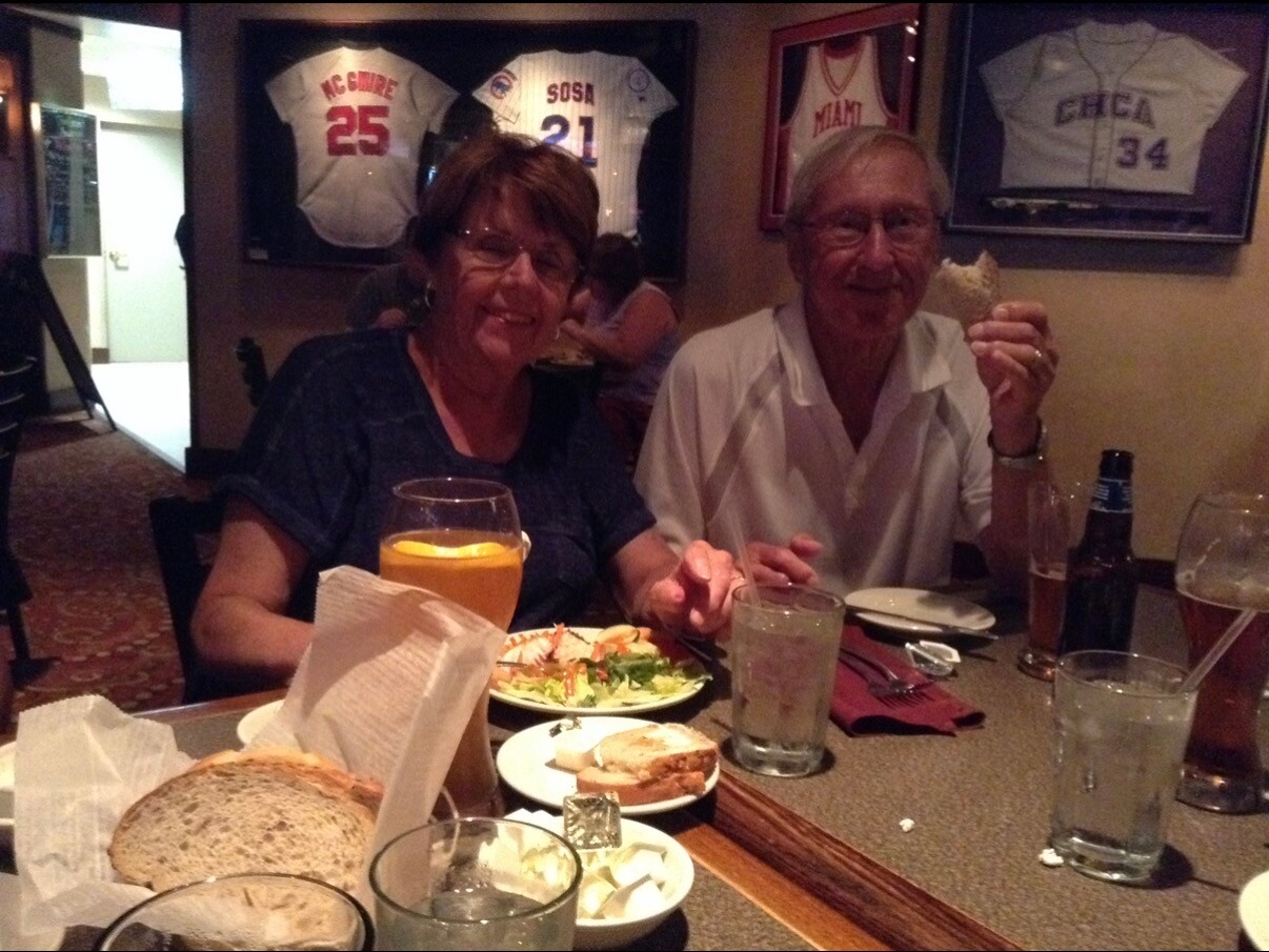 Two older adults dining together at a restaurant, smiling over a shared meal with bread, salad, and drinks on the table.