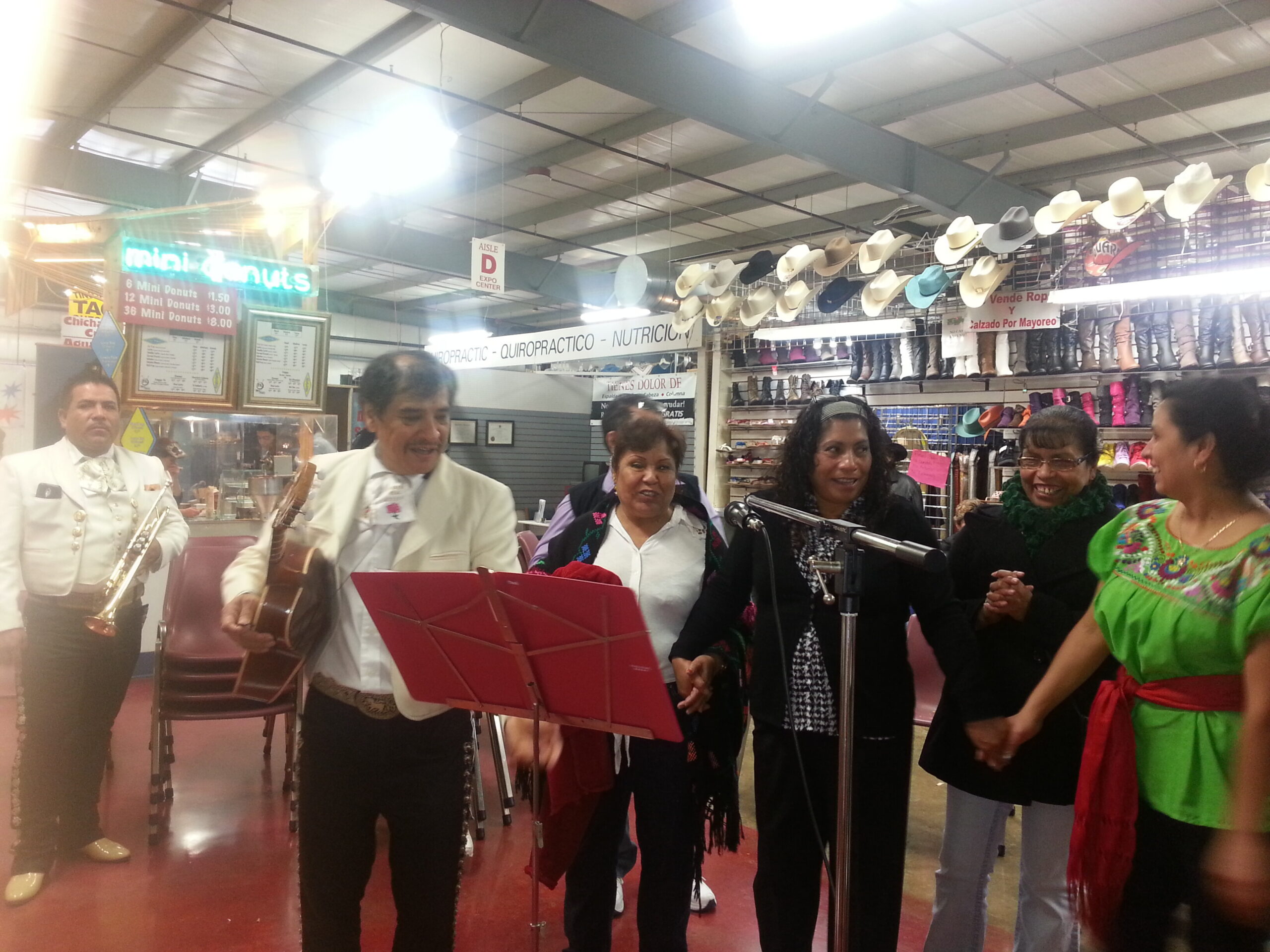 Mariachi band performing in a market booth with guitars, while shoppers watch; hats line the back wall.